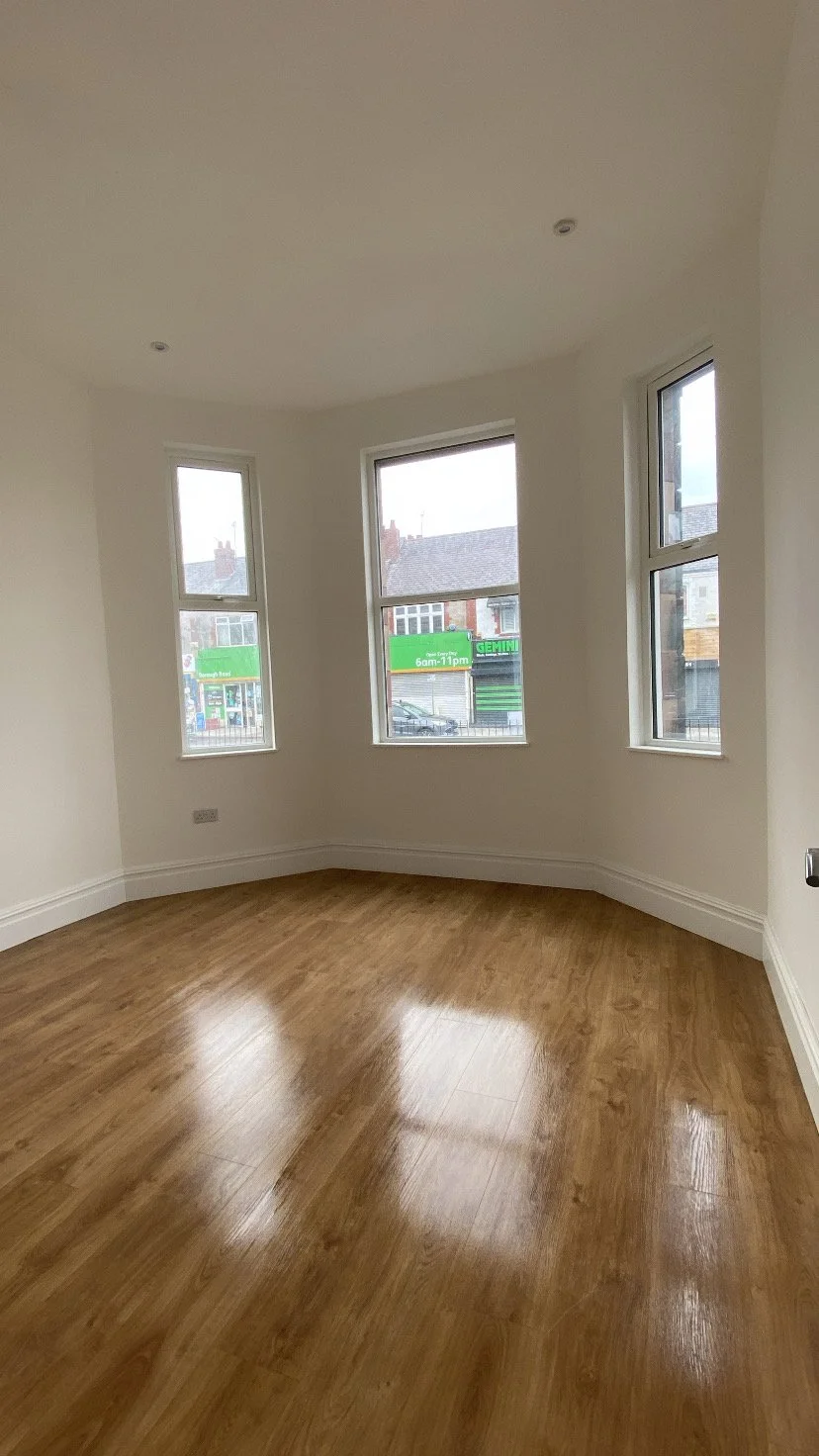 Empty room with white walls, wood floor, and three large windows showing a street view outside.