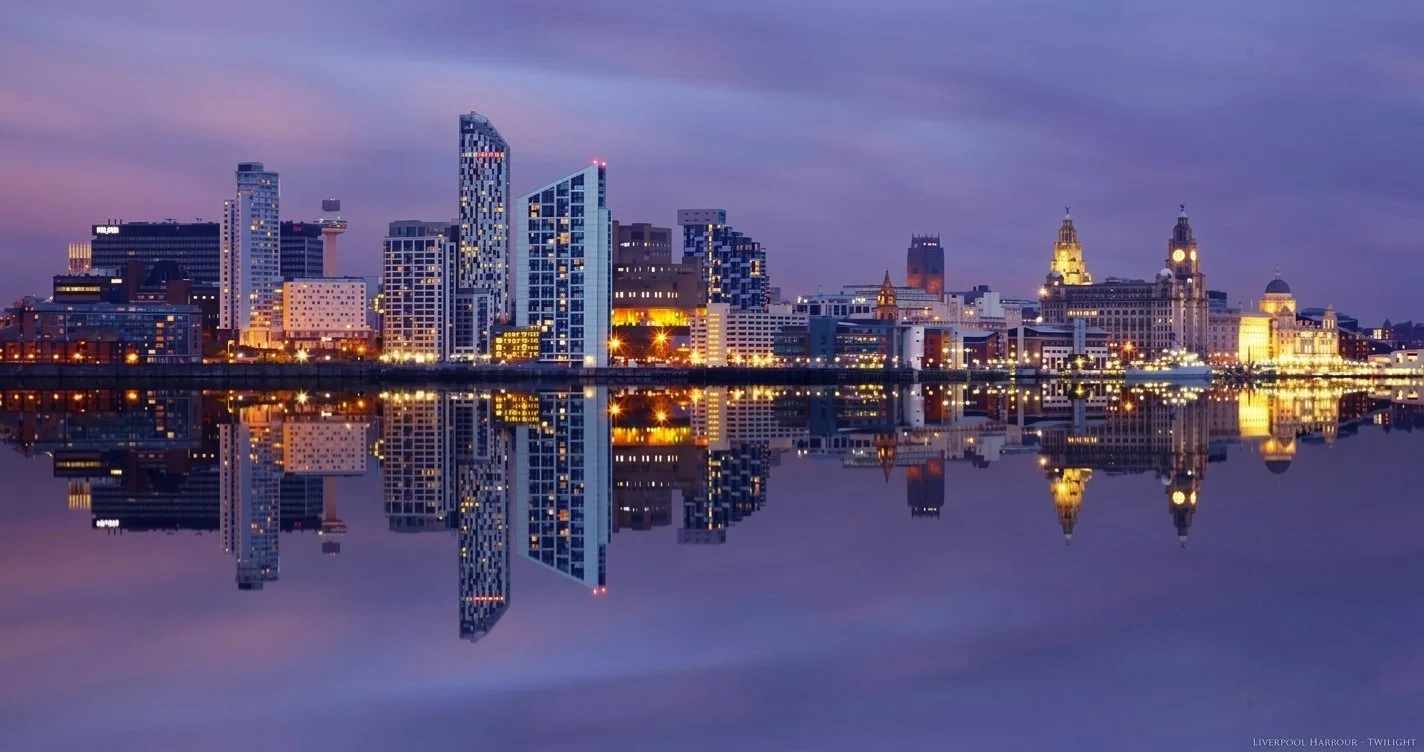 City skyline at twilight reflected in water, with modern high-rise buildings and historic structures, illuminated with lights.