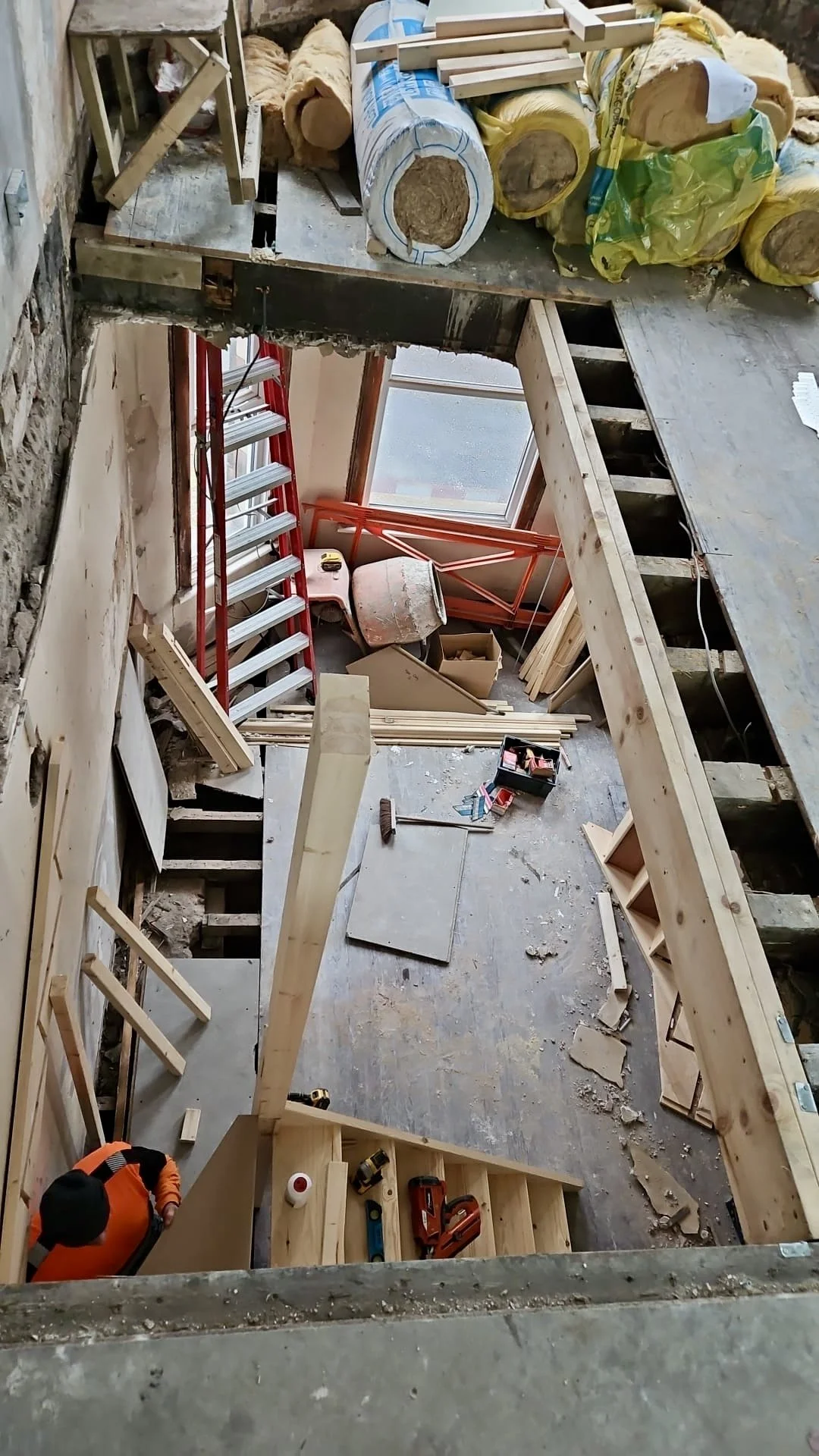 Construction site with unfinished wooden framing, a red ladder next to a large window, scattered tools, wood pieces, insulation, and construction materials.