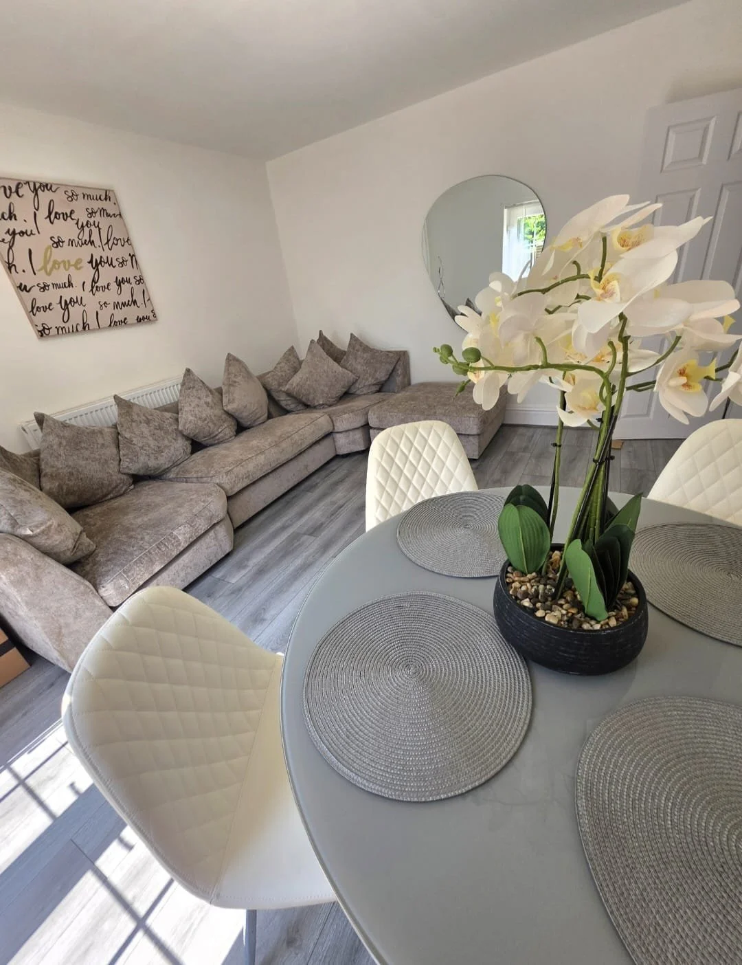 A living room with a beige sectional sofa, light-colored walls, a round mirror on the wall, a painting with black and gold script, a dining table with four white quilted chairs, and a centerpiece of white orchids in a black pot.