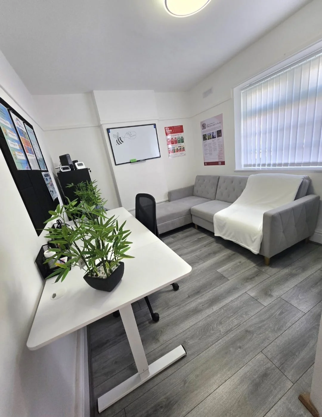 An office lounge with a white desk, a black office chair, a potted green plant, a gray sectional sofa with a white cover, a whiteboard with a bee drawing, and a window with vertical blinds.