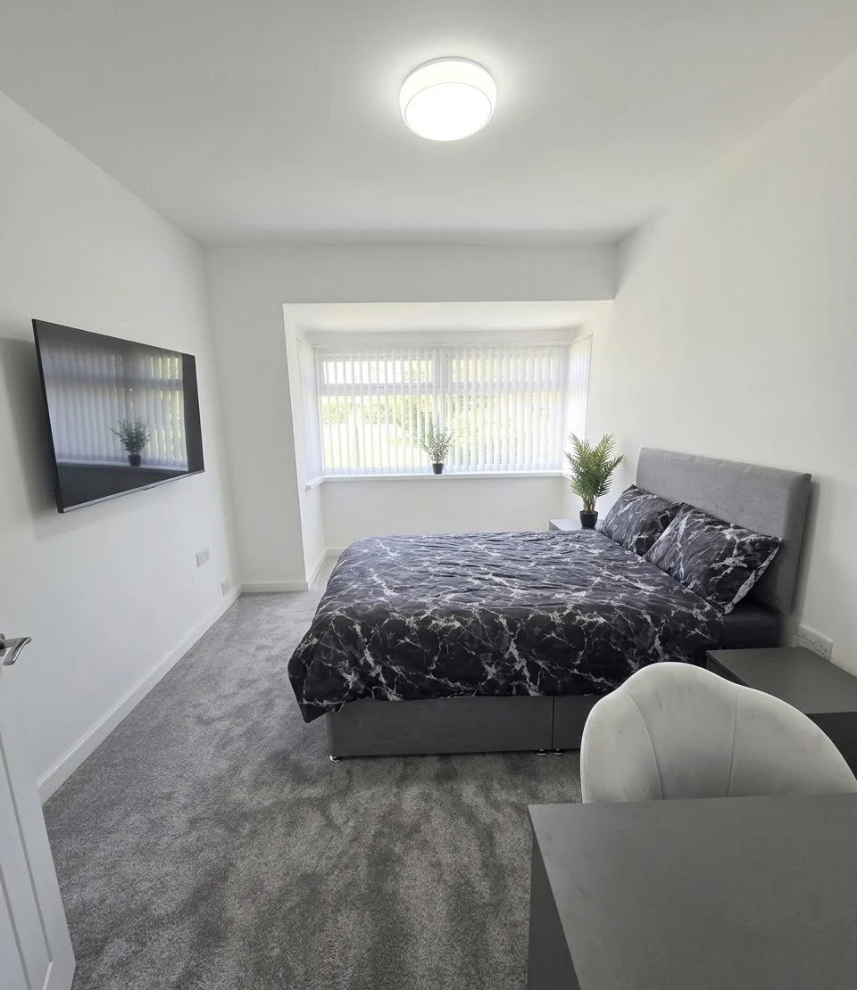A minimalist bedroom with a gray bed, black marble-patterned bedding, a gray headboard, and two small plants on the window sill. A flat-screen TV is mounted on the wall opposite the bed, and there is a white chair at a small black desk in the foreground. The room has a large window with vertical blinds, white walls, and a ceiling light.