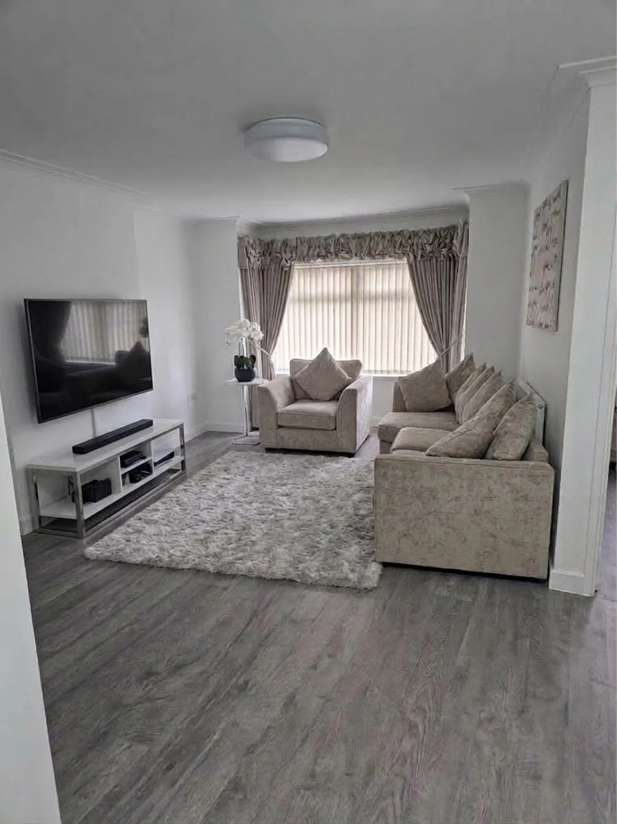 Living room with beige sofas, a TV on a stand, a window with pink curtains, a cream-colored shag rug, and a light-colored wood floor.