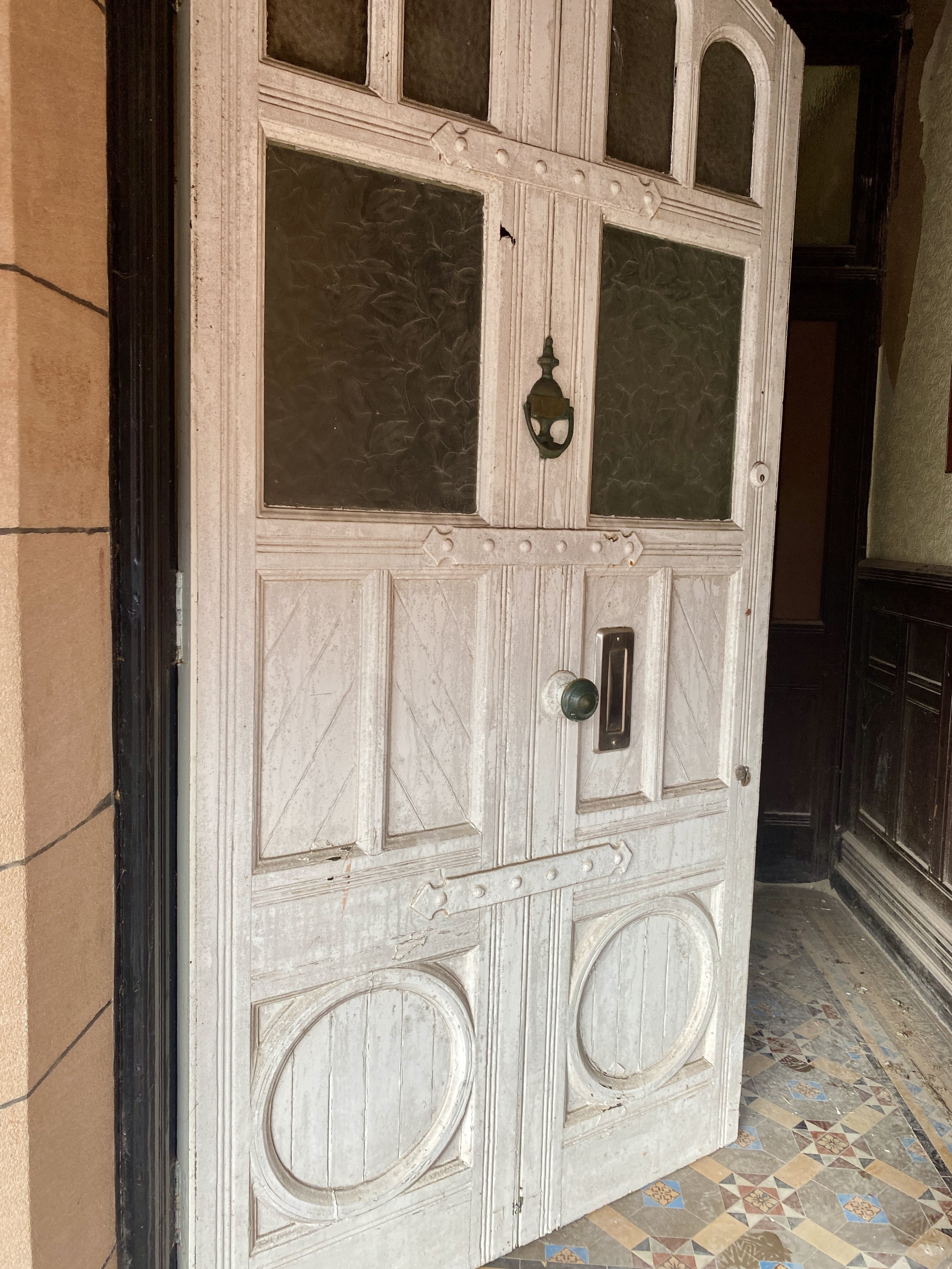 An old white wooden door with glass panels, a knocker, and a mail slot, set in a tiled entranceway.