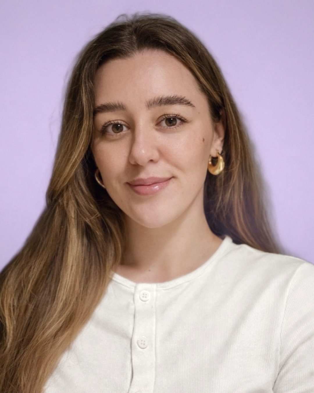A woman with long brown hair, wearing gold hoop earrings and a white button-up shirt, smiling against a light purple background.