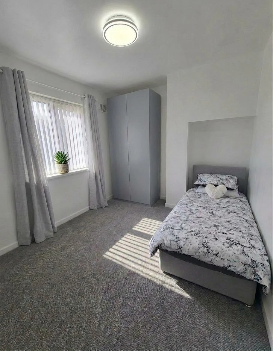 A small bedroom with gray walls, a single bed with marble-patterned bedding and a heart-shaped pillow, a gray wardrobe, a window with vertical blinds and light gray curtains, and a potted plant on the windowsill.