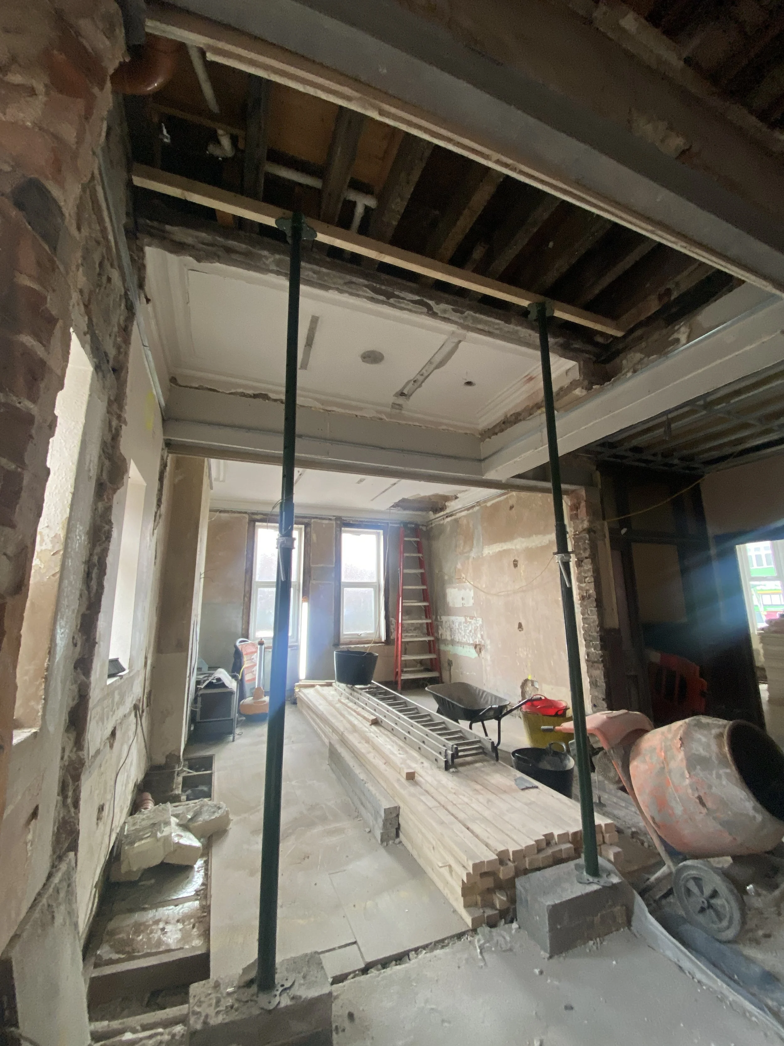 Room under renovation with exposed brick walls, ceiling open with wooden beams, construction tools, a ladder, buckets, and support poles.