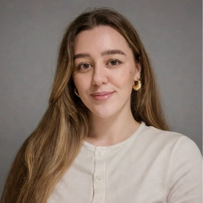 A young woman with long brown hair, wearing a white top and gold earrings, smiling softly at the camera against a plain gray background.
