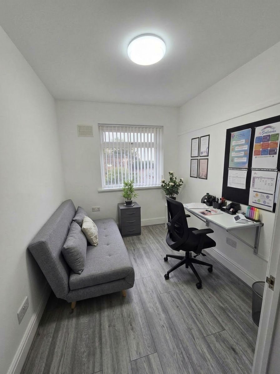 A small office with a gray sofa, a black office chair, a white desk with office supplies, potted plants, framed certificates on the wall, and a window with vertical blinds.