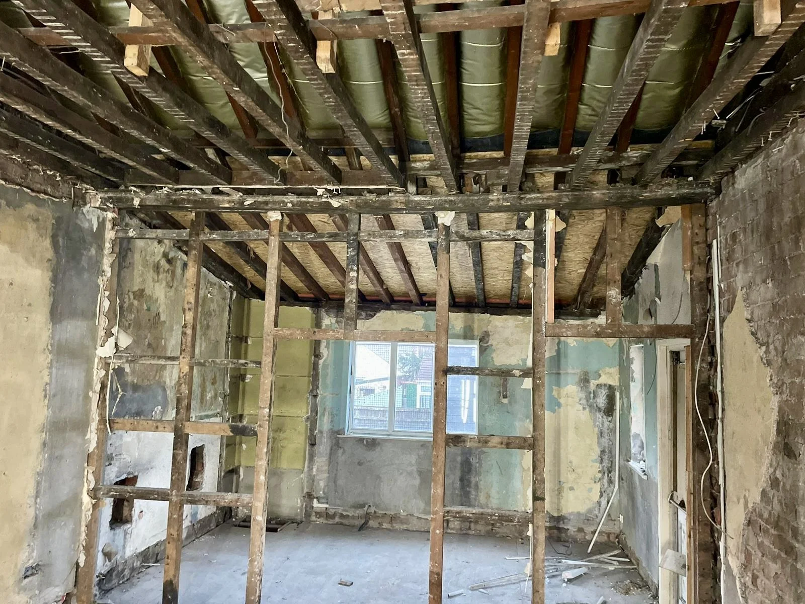 Interior of a house under renovation with exposed wooden framing, walls in disrepair, and a window providing natural light.