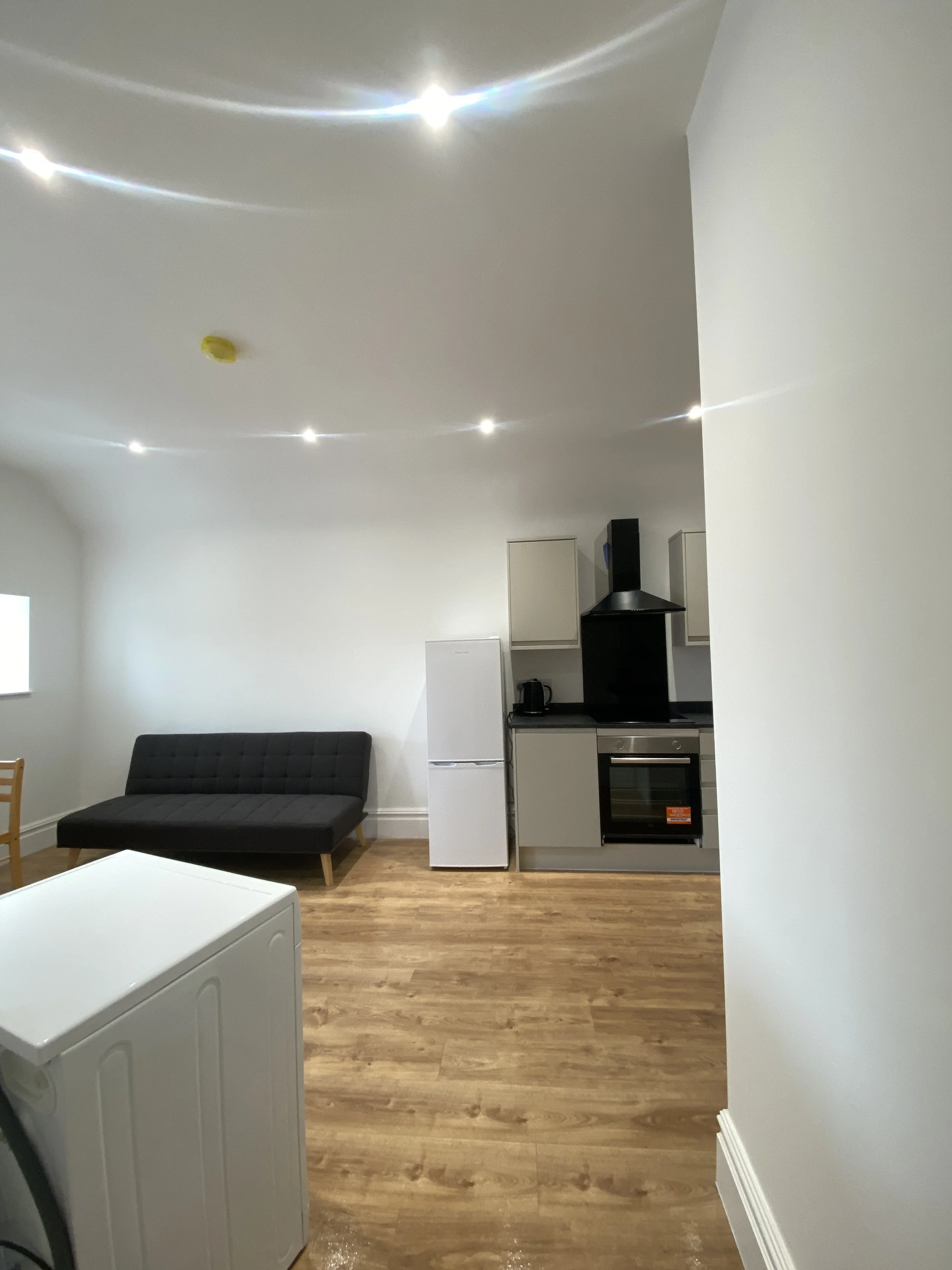 Interior view of a modern kitchen with white walls, a black sofa, a white refrigerator, and a built-in oven. The ceiling has multiple bright recessed lights, and the wood flooring adds warmth to the space.