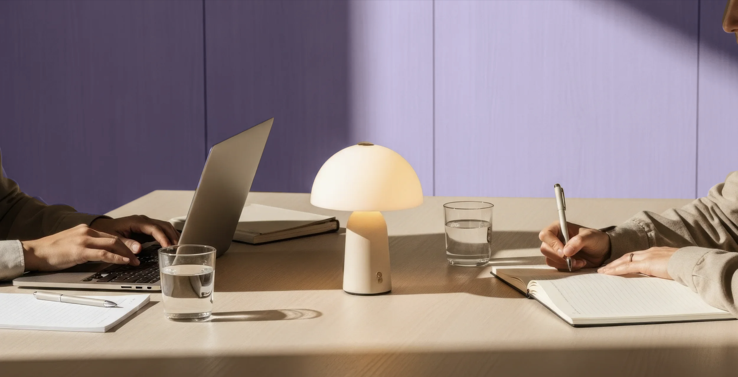 Two people sitting at a light wood table with notebooks, glasses of water, a laptop, a pen, and a small lamp between them, with purple wood-paneled wall in the background.