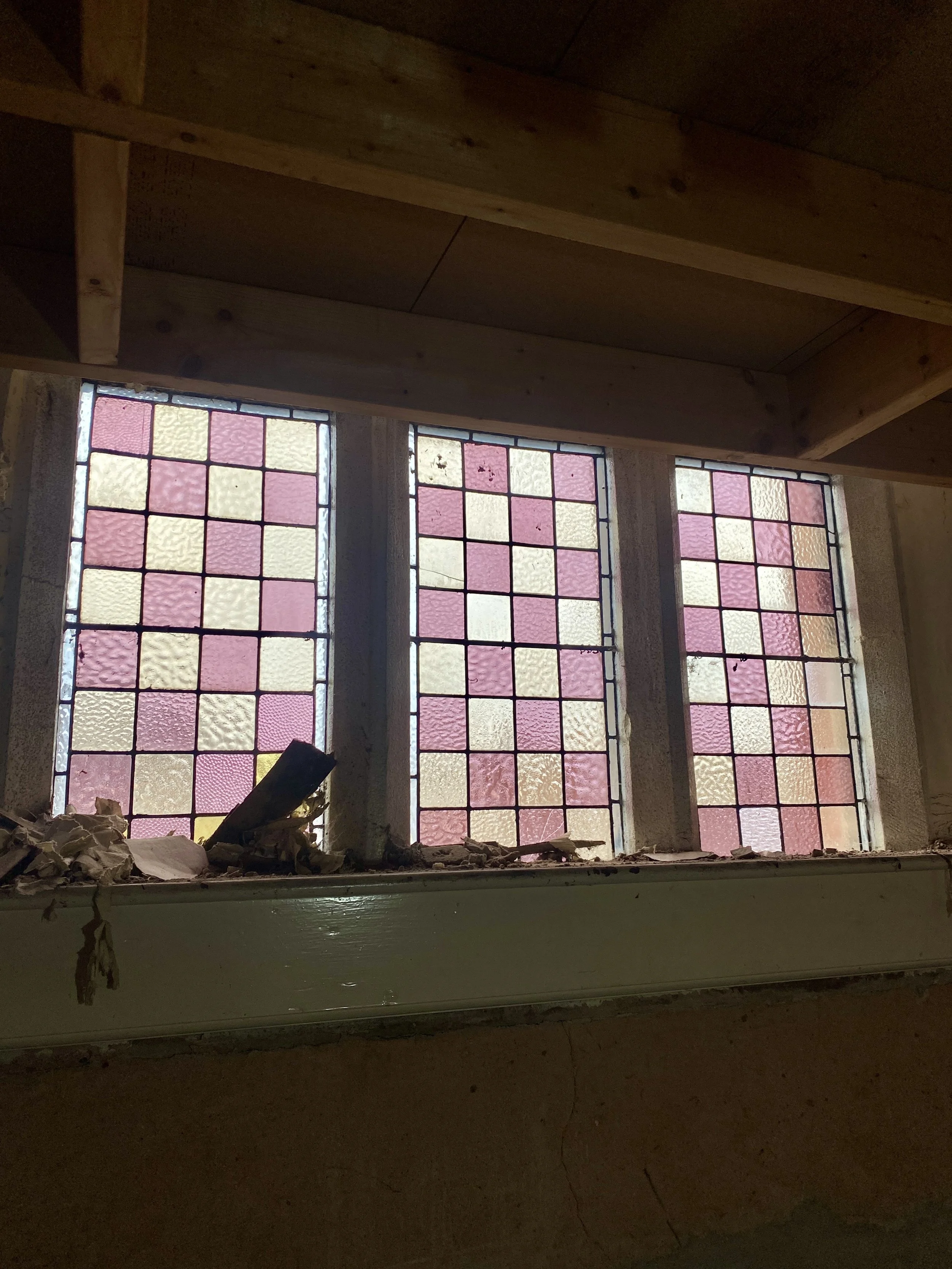 The image shows stained glass windows with a pattern of pink, yellow, and orange squares, viewed from inside a room under construction or renovation, with debris and insulation material near the window sill.