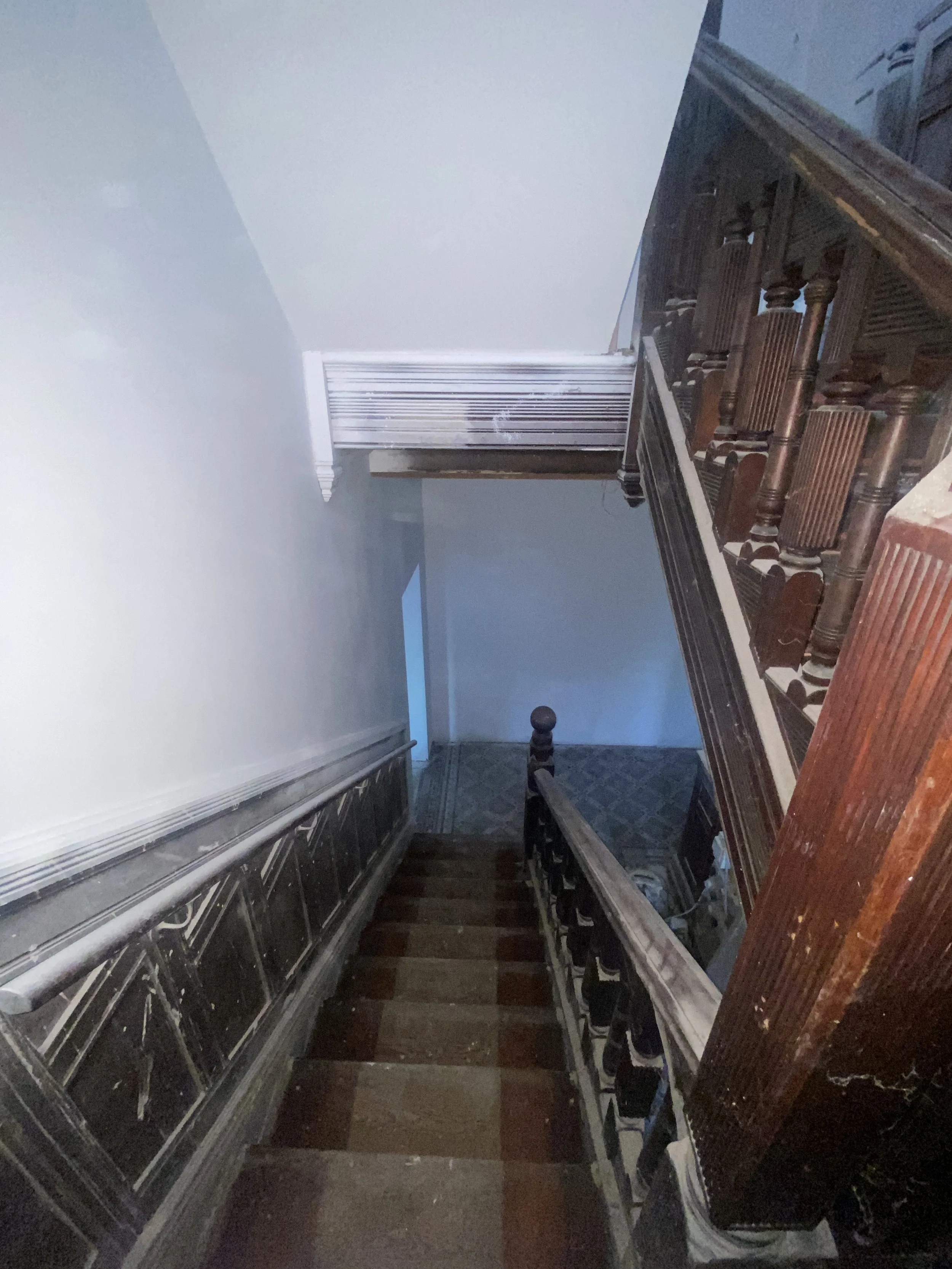 A dimly lit staircase with wooden handrails and black metal balusters, leading down to a lower level with tiled flooring, next to a white wall with a vent and a doorframe.