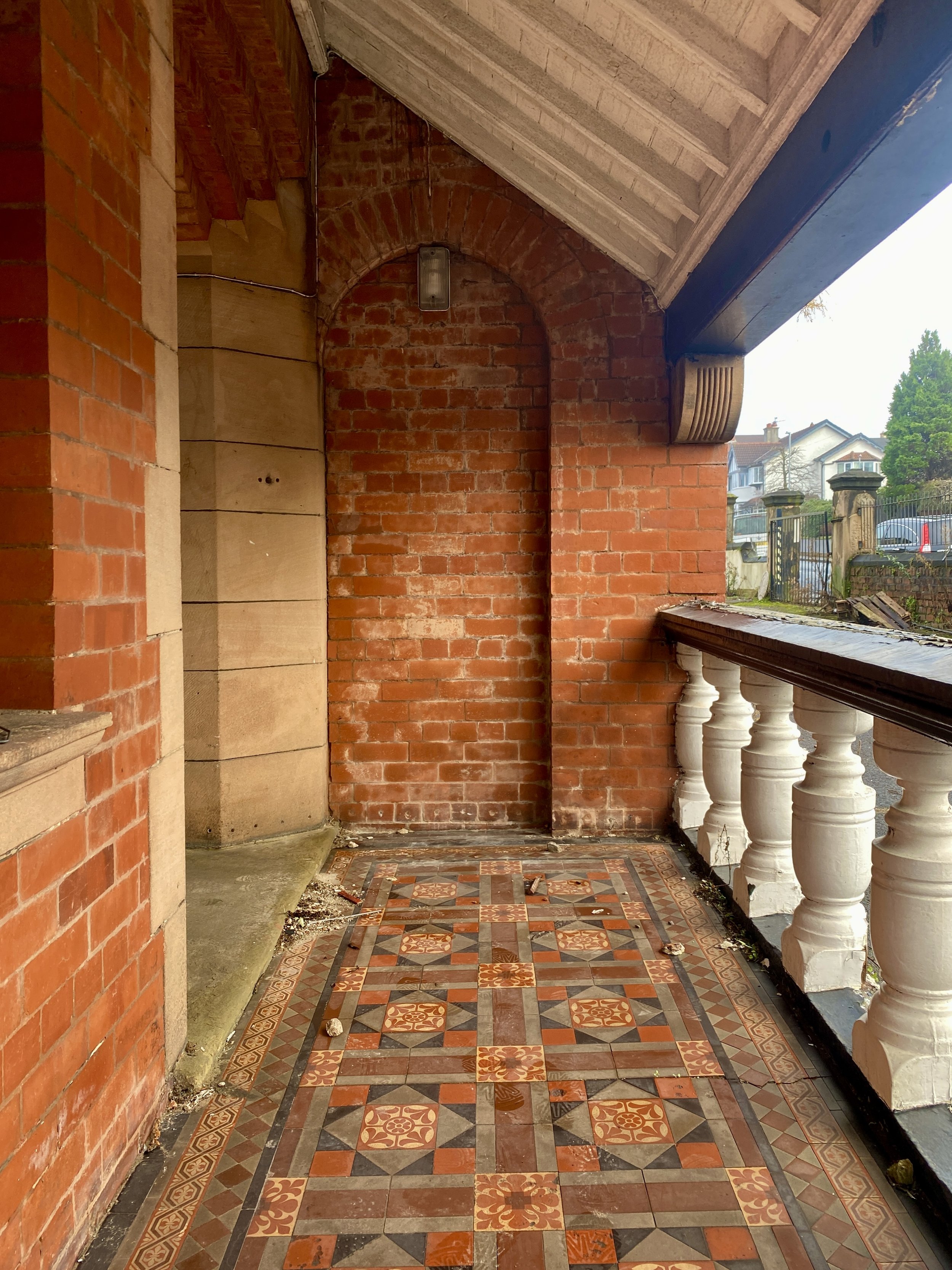 Porch with brick wall and ornate tiled floor, white balustrade on right, brick archway to the left, outside view of houses and trees in the background.