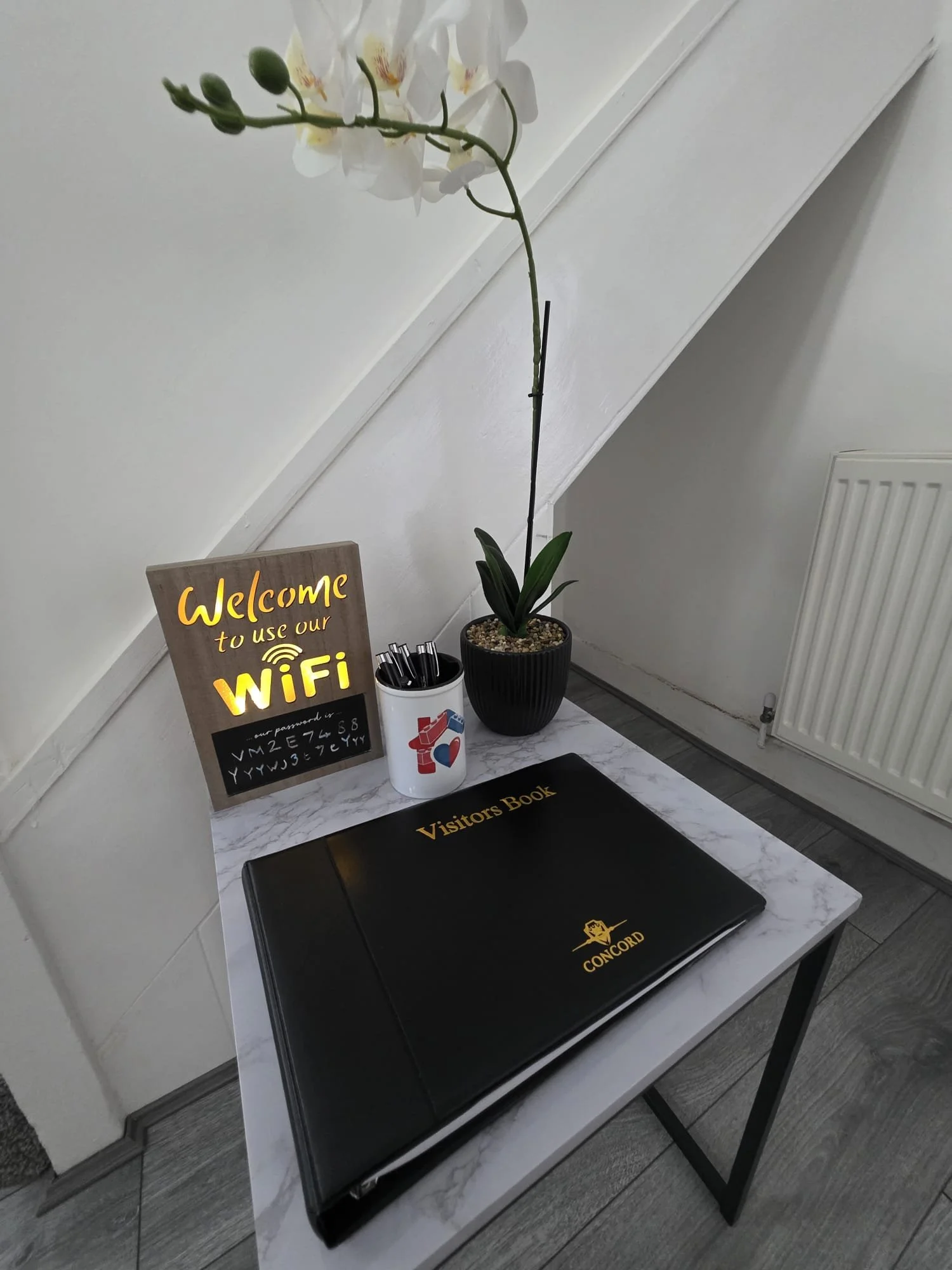A small table with a black Concord laptop, a visitors book, a white cup with black markers, an orchid plant, and a sign that says 'Welcome to use our Wi-Fi' inside a room with white walls and a radiator.