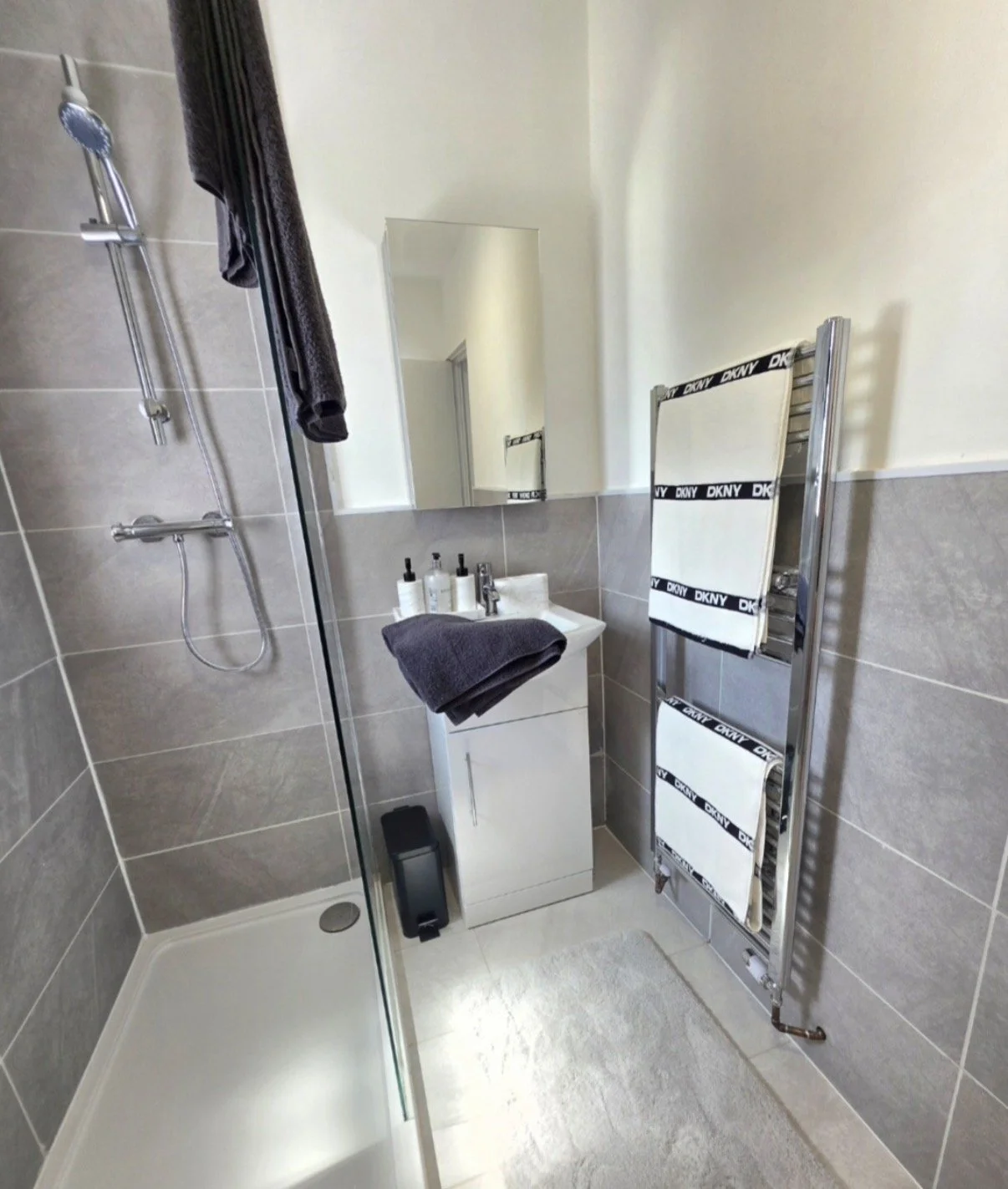 Small modern bathroom with a walk-in shower, gray tiled walls, a white sink with black towels and toiletries, a mirror, and a heated towel rack with DKNY towels.