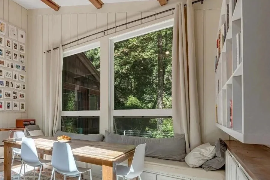Bright dining area with large windows overlooking greenery, white walls, a wooden table with white chairs, a built-in bench with cushions, and a wall with photo frames and shelves filled with books. Bayly Builds window installation.
