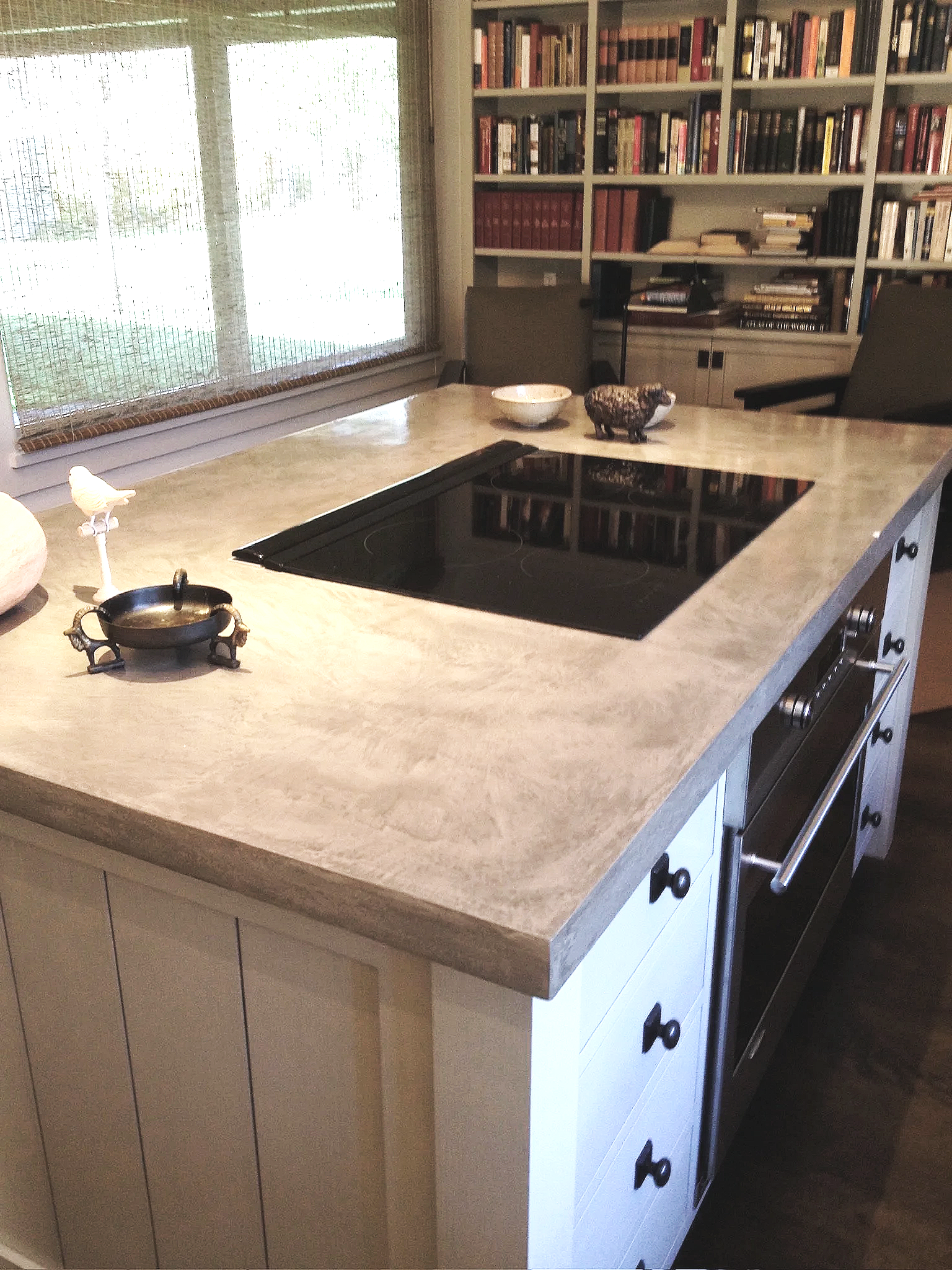 A kitchen with white cabinets, a concre countertop, and black stove top. Behind the counter is a window with blinds and a large white bookshelf filled with books. Bayly Builds Concrete Countertop Installation.