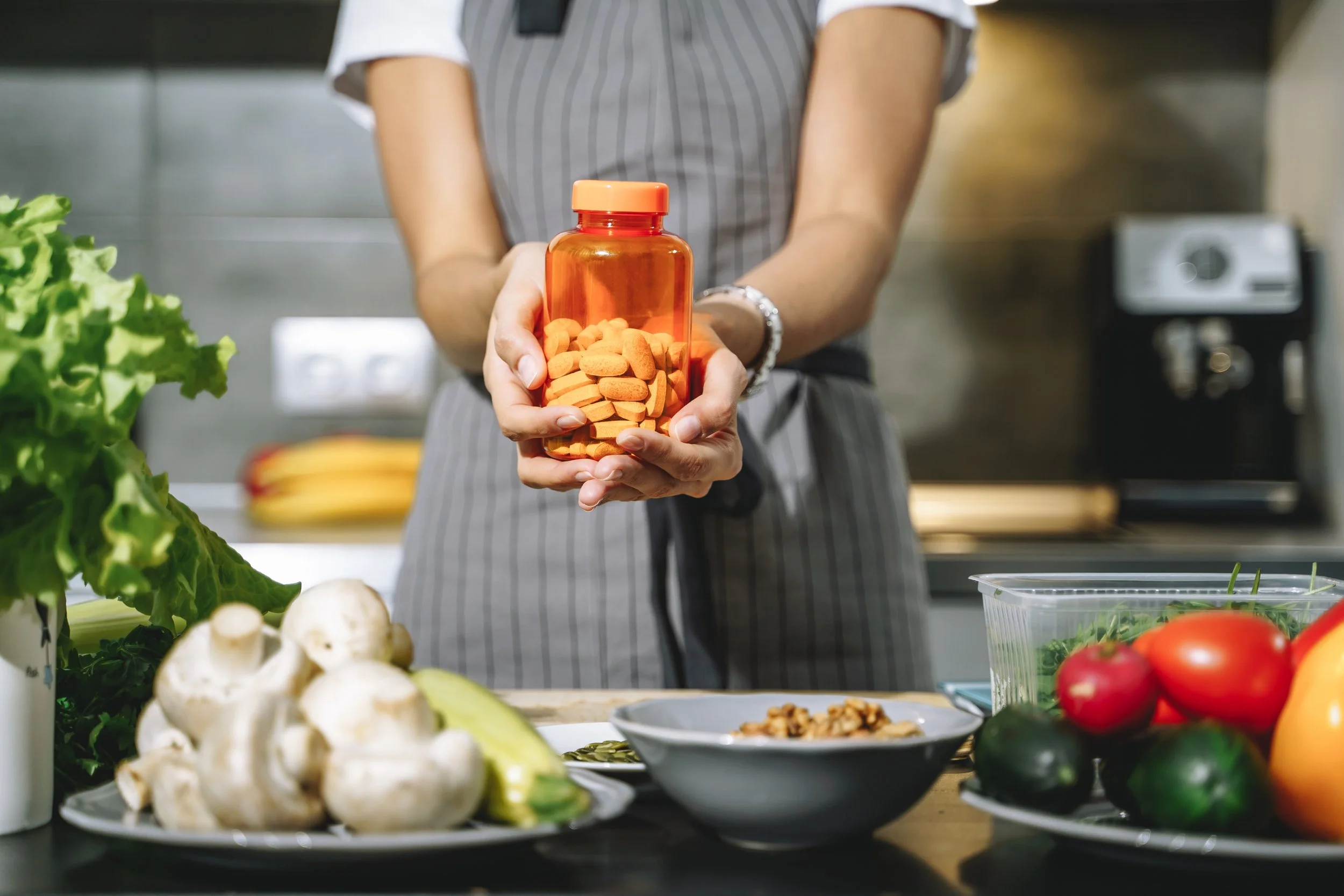 Person holding an orange pill bottle filled with pills in a kitchen or food preparation area surrounded by fresh vegetables and ingredients.