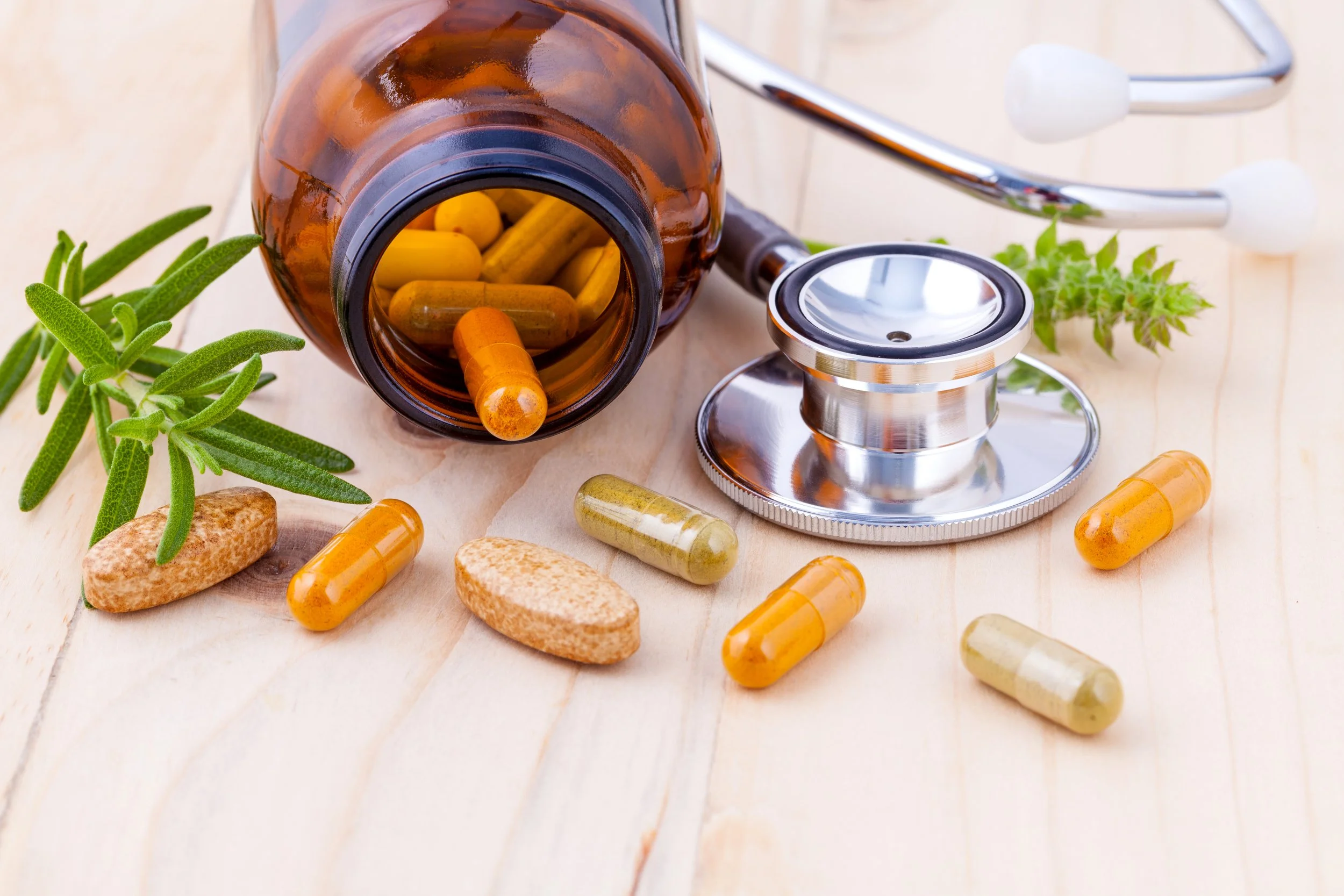 An open brown pill bottle spilling yellow, green, and beige capsules onto a light wooden surface, with a stethoscope and sprigs of green herbs nearby.