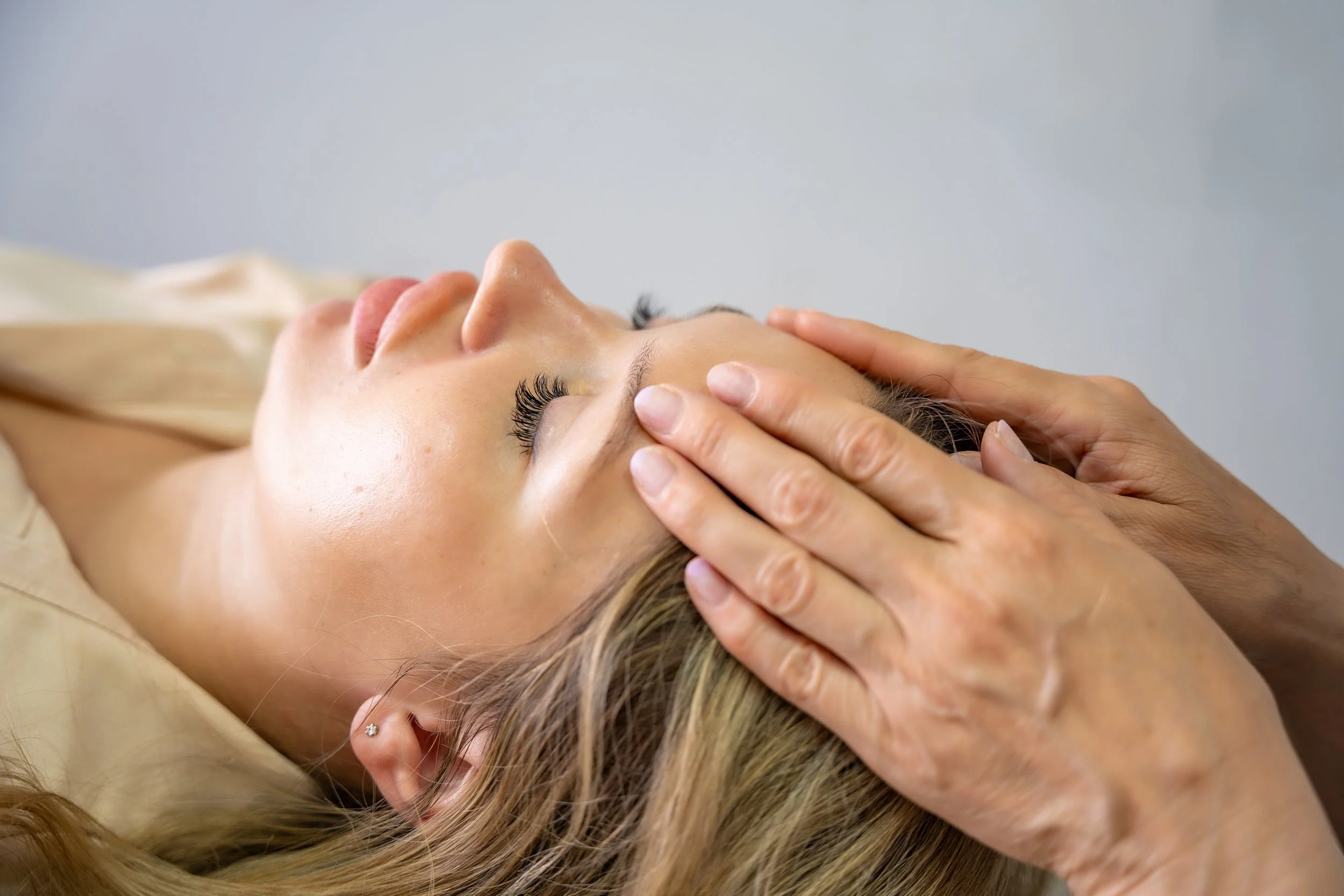 A woman lying down with her eyes closed receives a facial massage from a professional massage therapist.