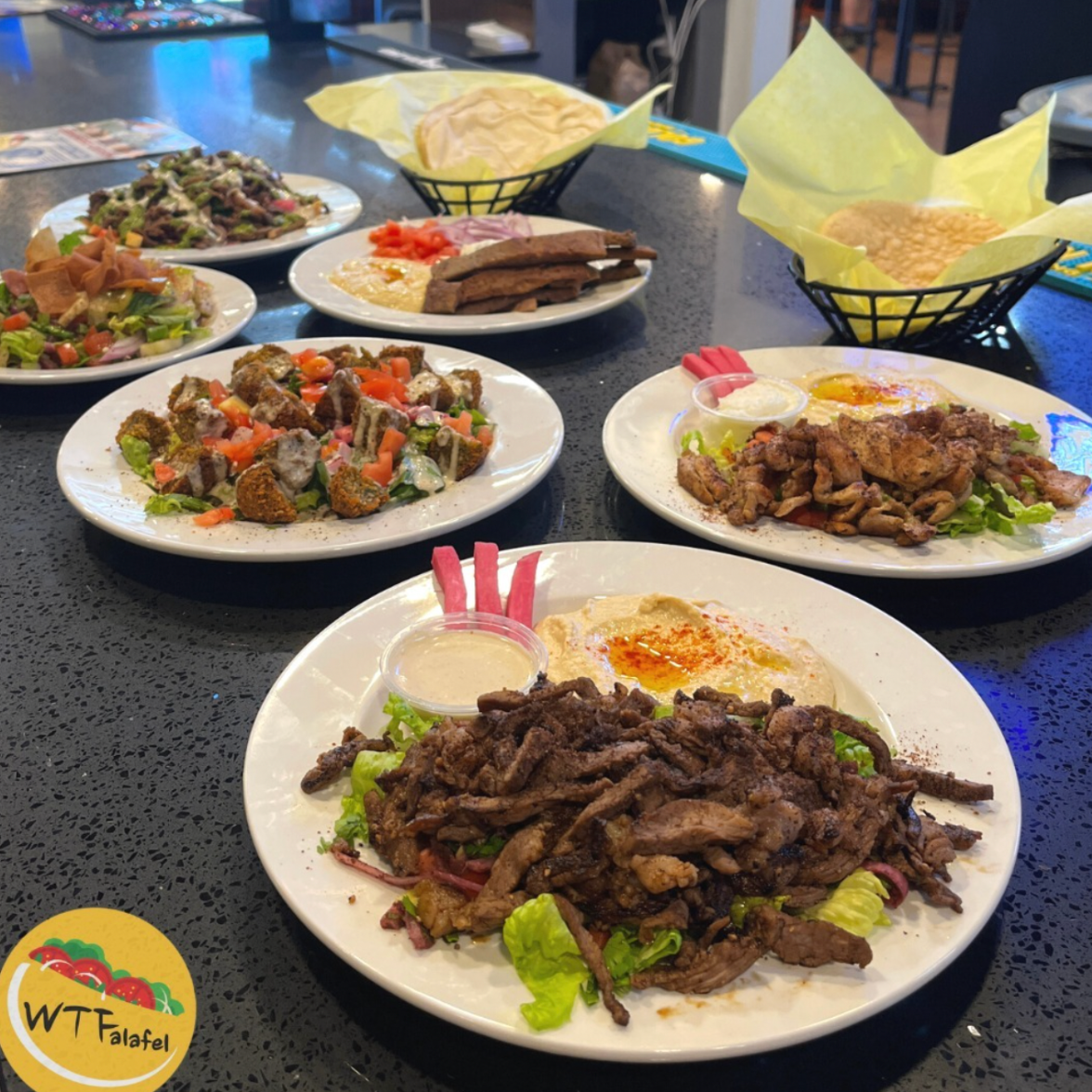 Assorted Middle Eastern dishes on white plates, including grilled meats, fried vegetables, salads, hummus, pita bread, and dipping sauces, on a black speckled table.