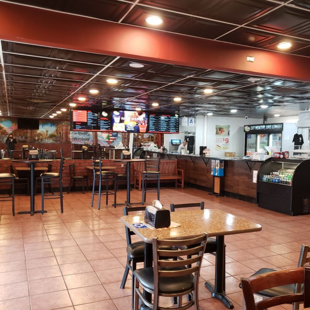 Empty restaurant with a few tables and chairs, a counter area with digital menu boards, and a mural on the wall in the background.