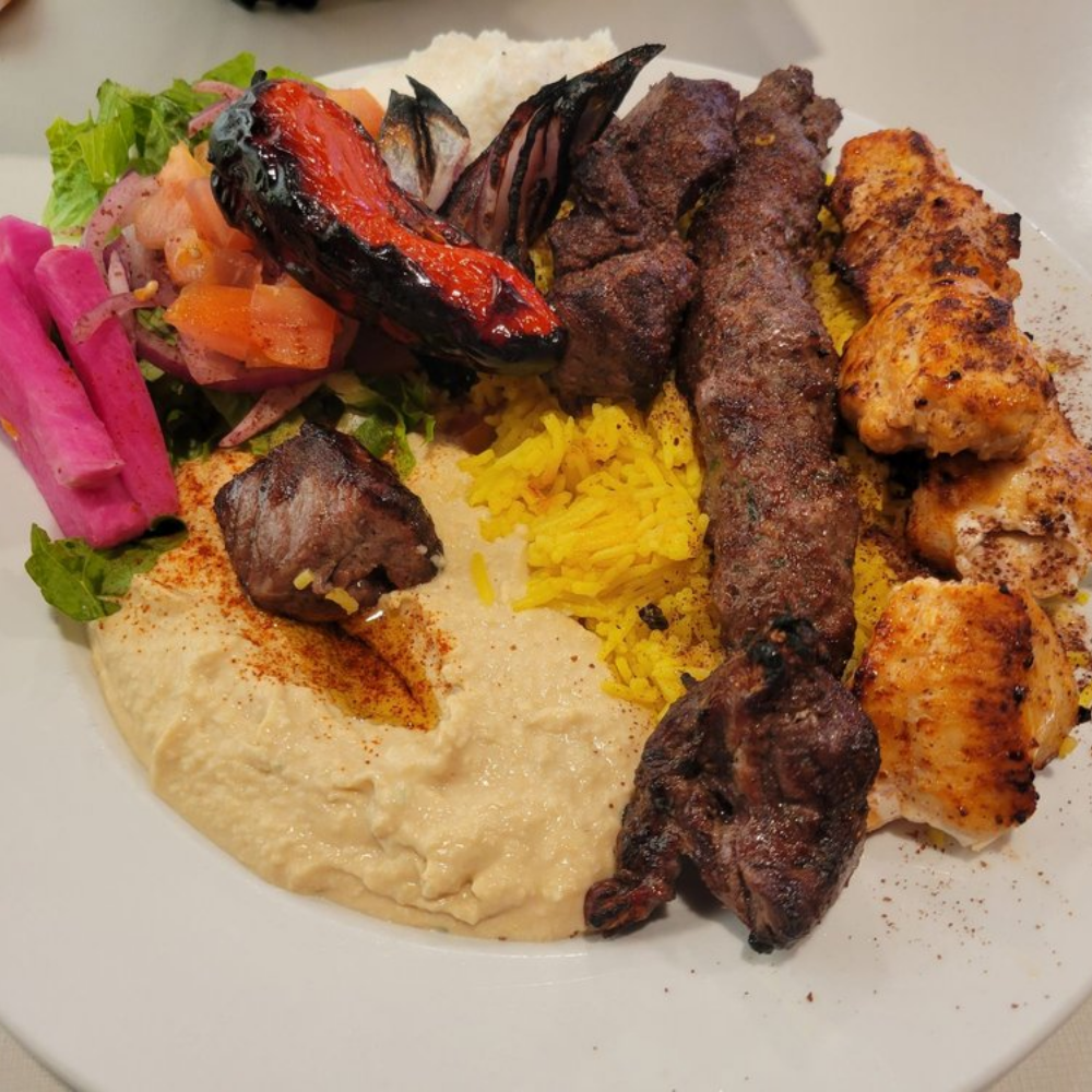 A plate of Middle Eastern food with grilled meats, rice, grilled vegetables, salad, hummus, and pickled vegetables.