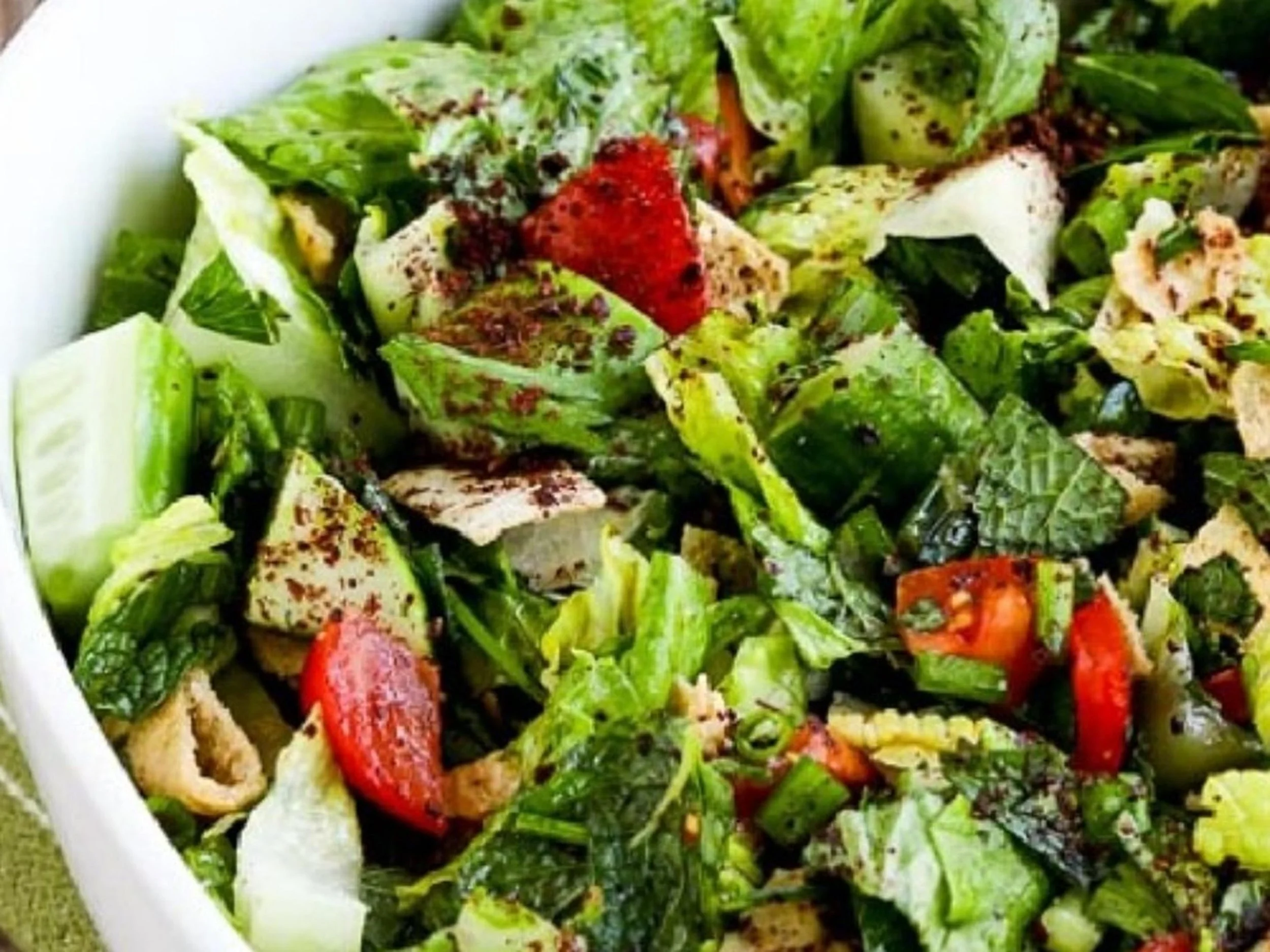Close-up of a fresh vegetable salad with chopped cucumbers, tomatoes, lettuce, and herbs in a white bowl.