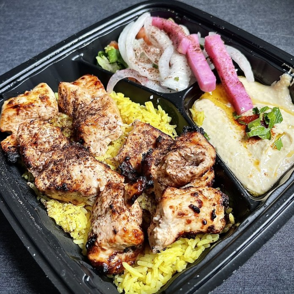 A black meal tray containing grilled chicken drumsticks on yellow rice, a side of potato salad with onions, and a serving of creamy mashed potatoes garnished with green herbs.