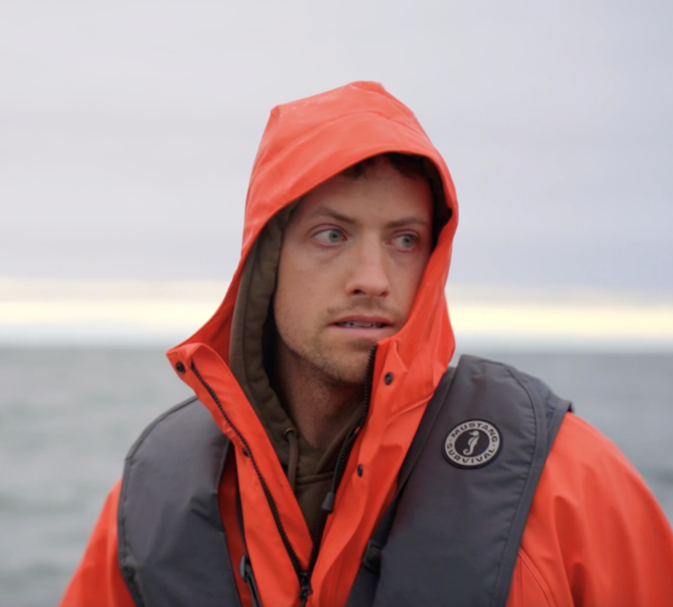 A man wearing an orange hooded rain jacket and a life vest outdoors near the water, looking to his left with a serious expression.