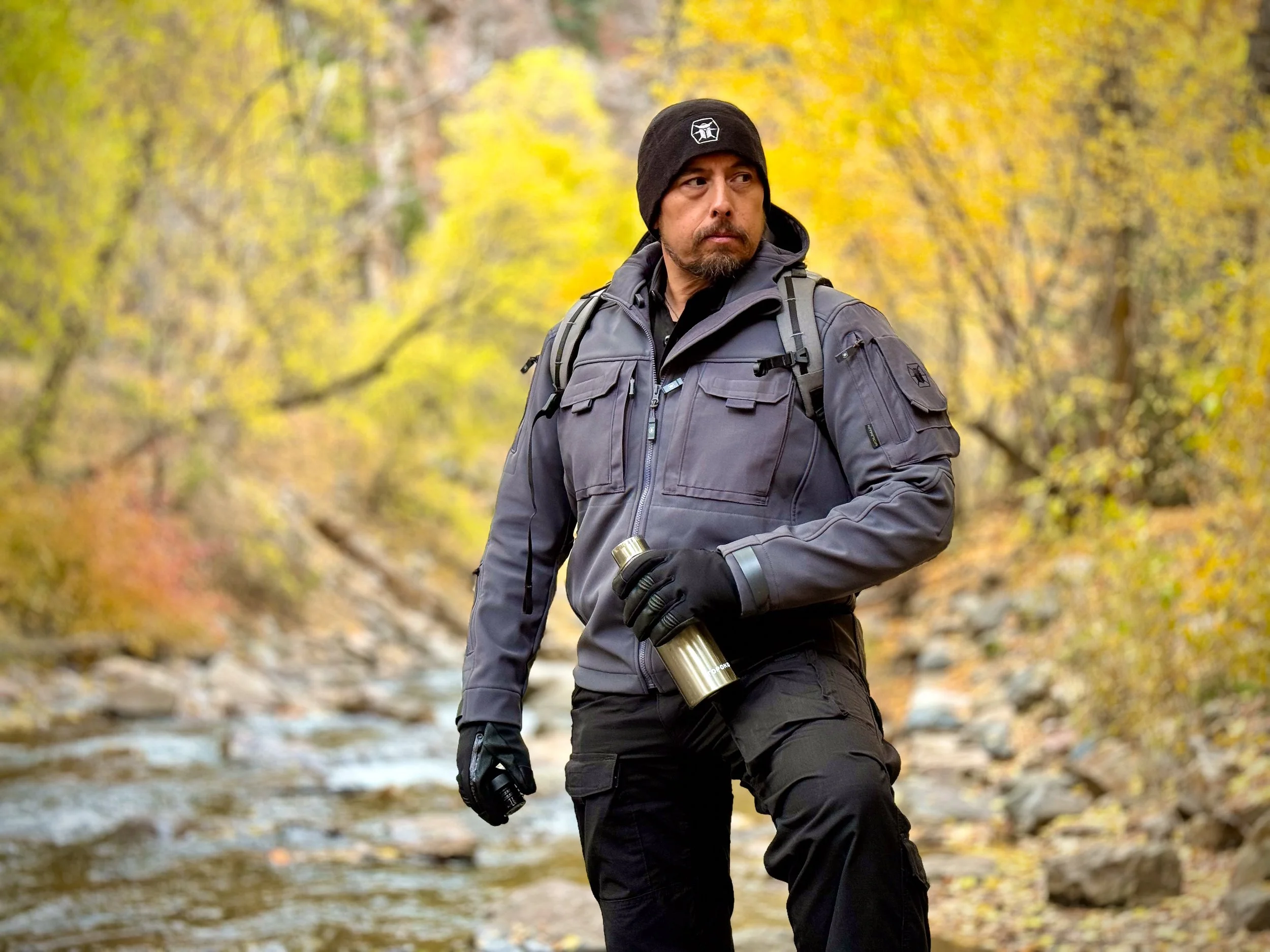 A man in outdoor hiking gear, including a black beanie with a logo, a gray jacket, and black gloves, walking in a forested area with yellow and orange autumn leaves and a creek.