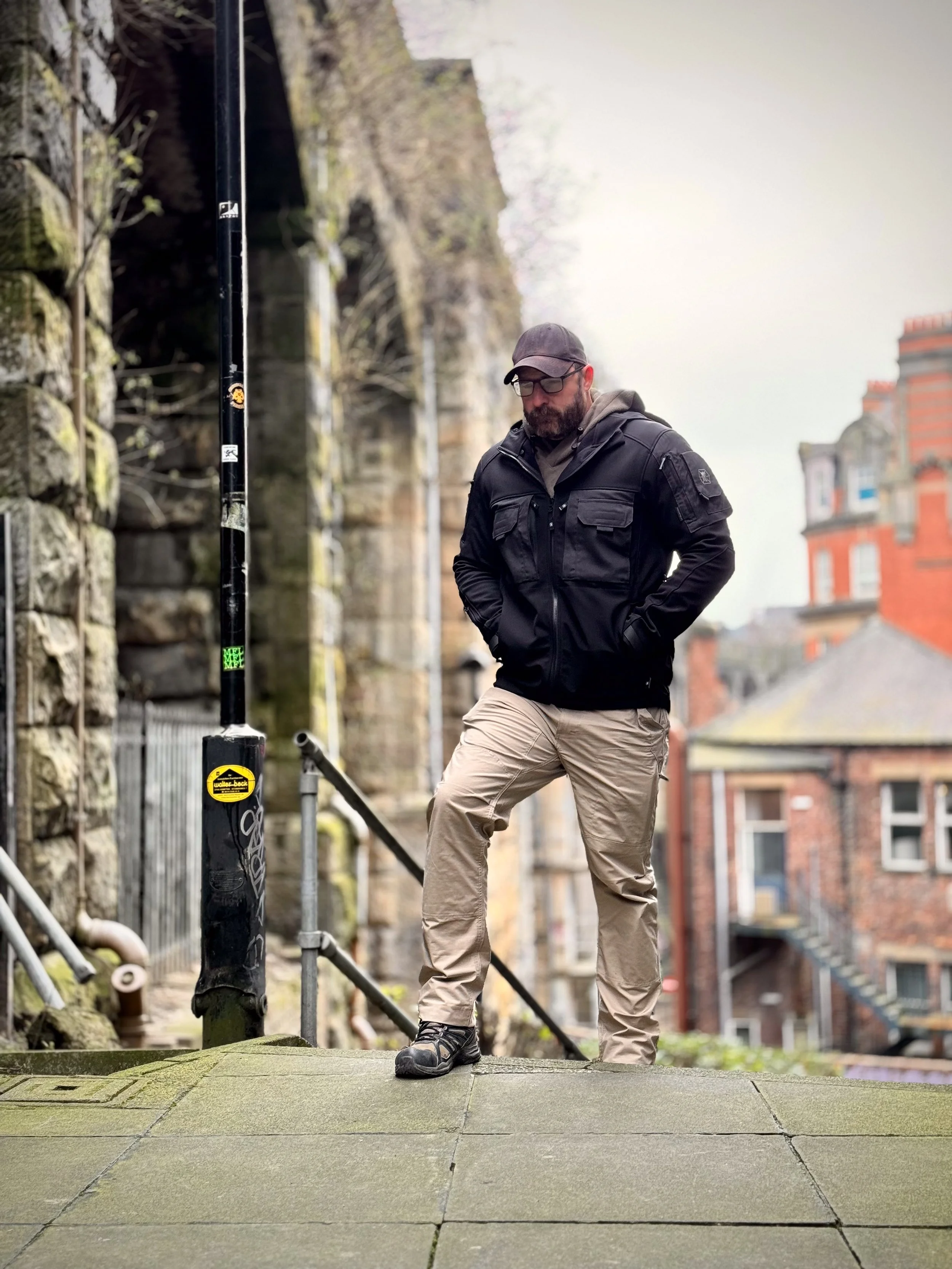 A man with a beard in outdoor clothing and glasses walks up a steep paved street past a black post with a yellow sticker, beside a stone wall and in front of brick buildings with various architectural details.