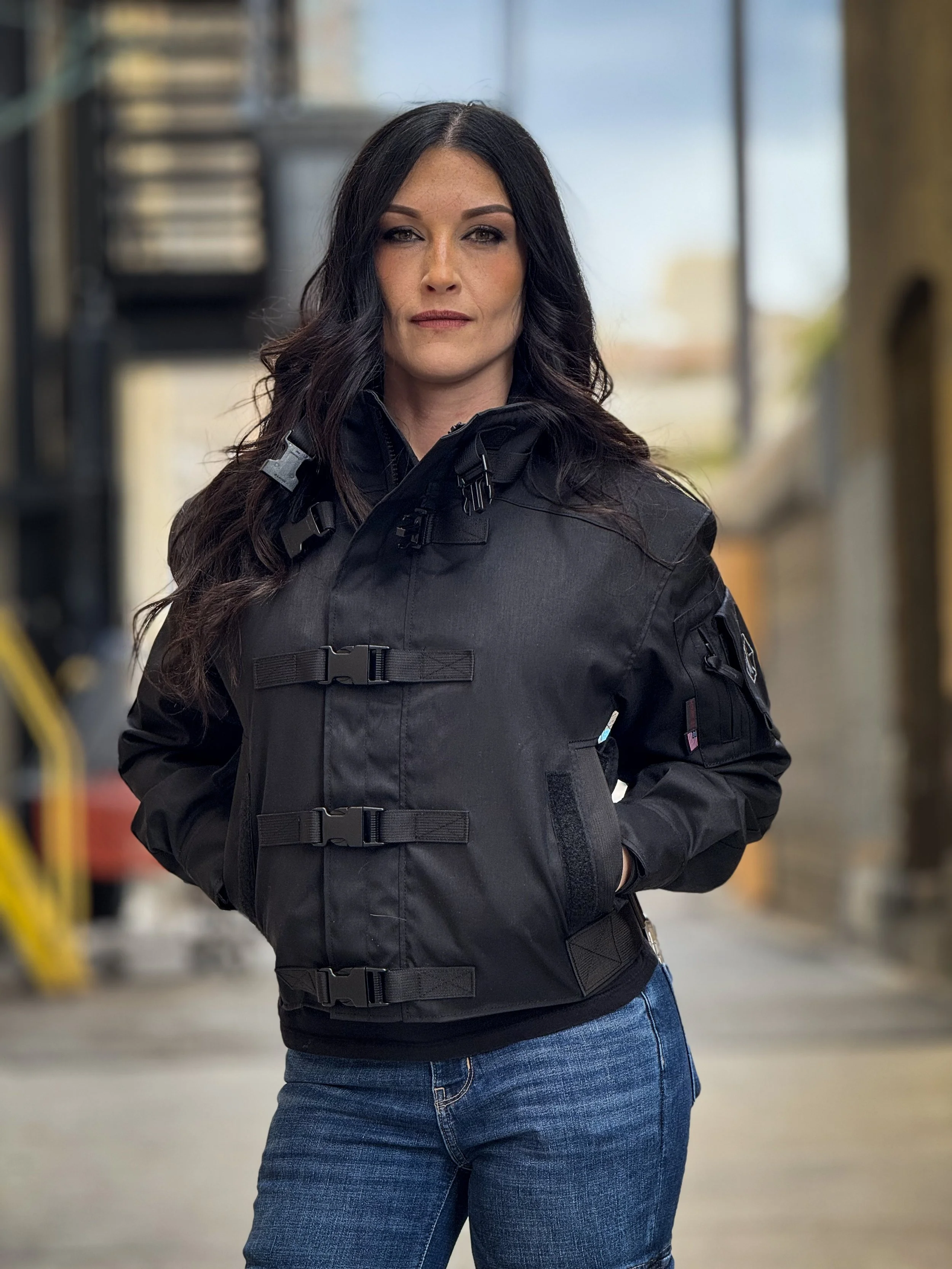 Woman with long dark hair wearing a black tactical jacket with buckles and blue jeans, standing indoors.