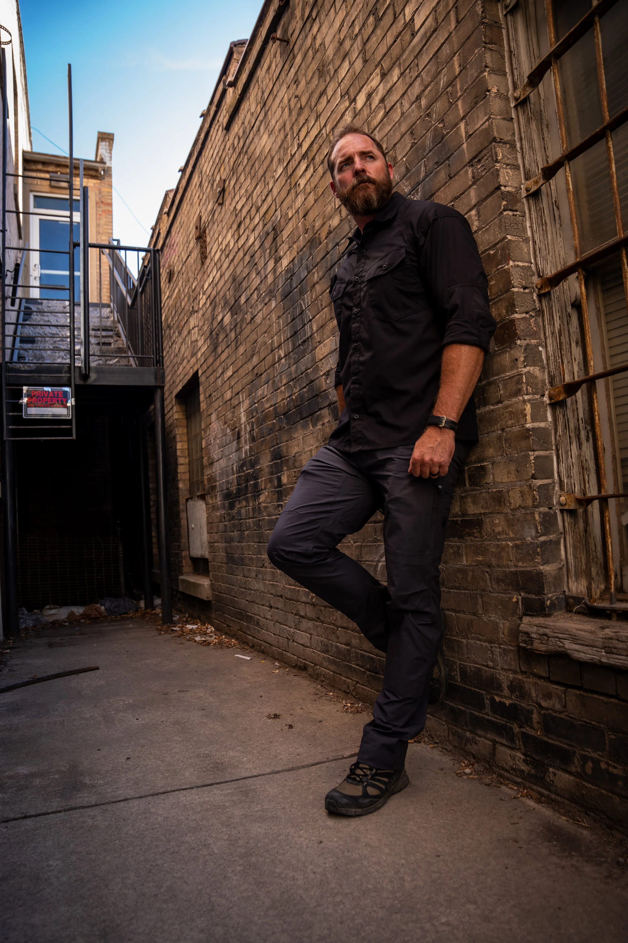 A man with a beard wearing a black shirt, gray pants, and hiking shoes leaning against a brick wall in an alleyway with a staircase and a sleeping sign.