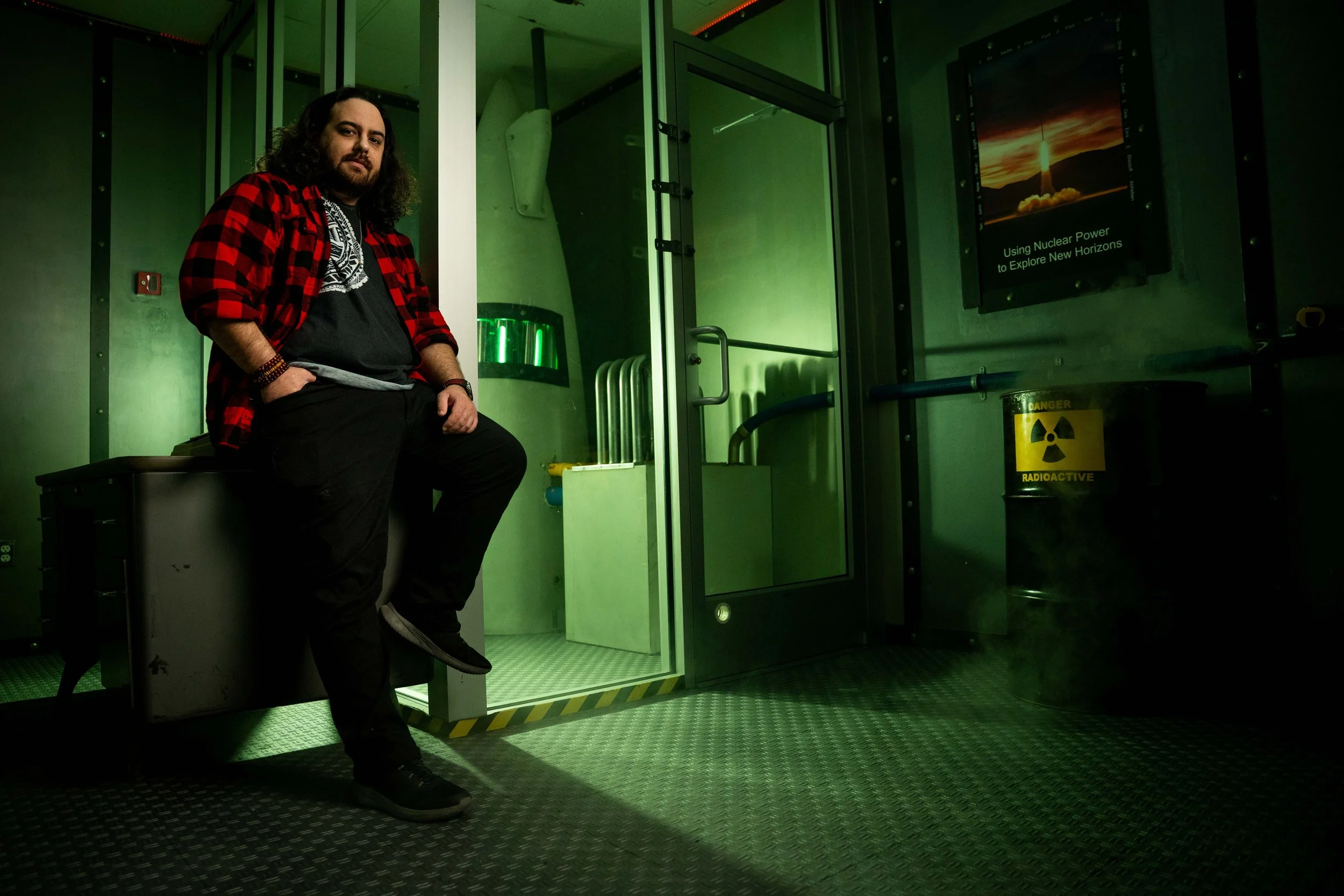 A man with long hair and a beard, wearing a red and black plaid shirt over a black graphic T-shirt, stands inside a green-lit room next to a radiation hazard barrel. The room has a glass door with a sign that says 'Using Nuclear Power to Explore New 