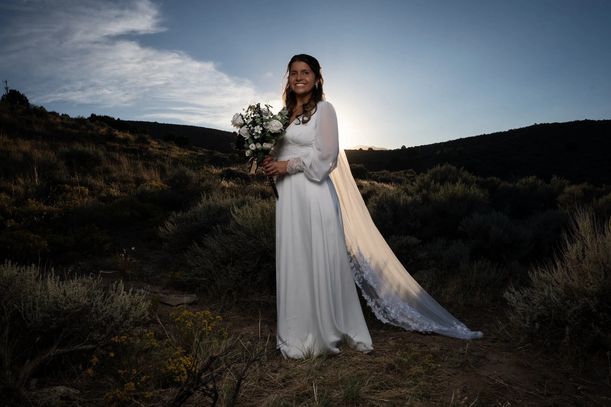 A woman in a white wedding dress holding a bouquet of white and green flowers, standing outdoors on a hillside during sunset or sunrise with a partly cloudy sky.