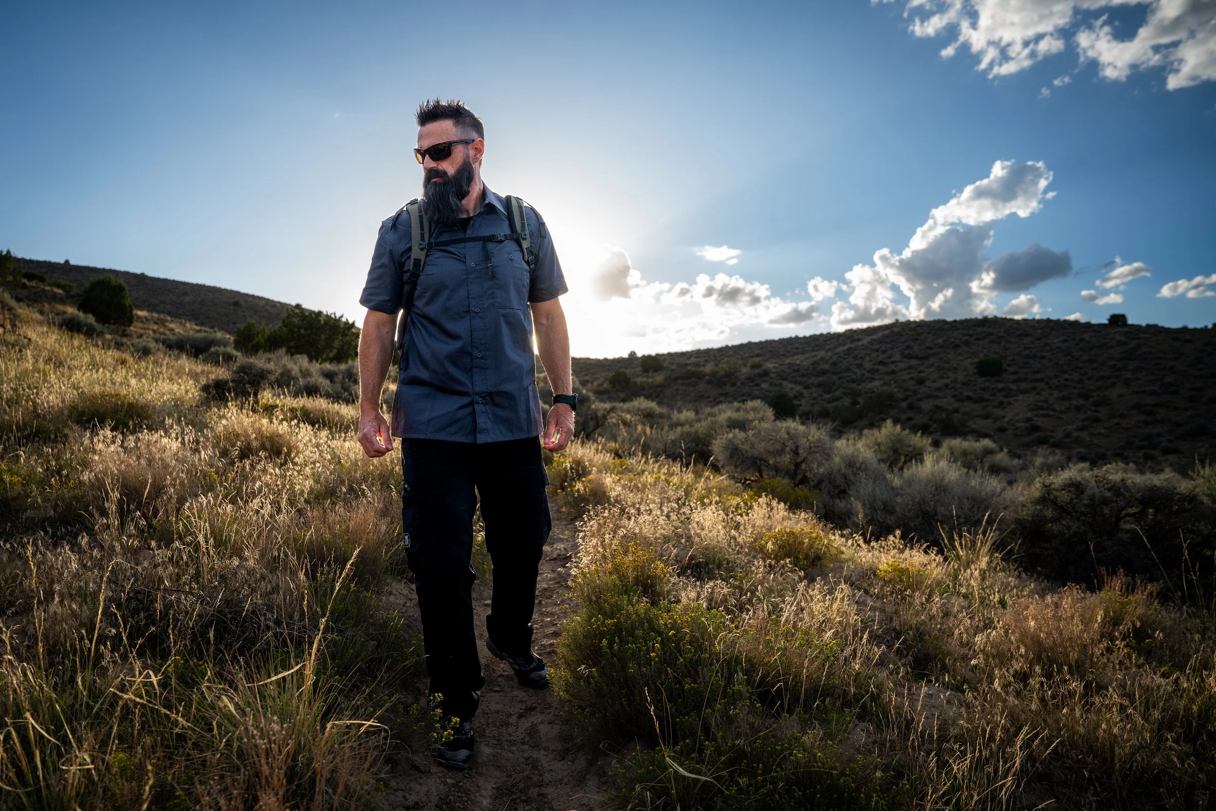 A man with a beard and sunglasses walking on a dirt trail through a grassy and shrub-covered hillside during sunset or sunrise, with a partly cloudy sky in the background.