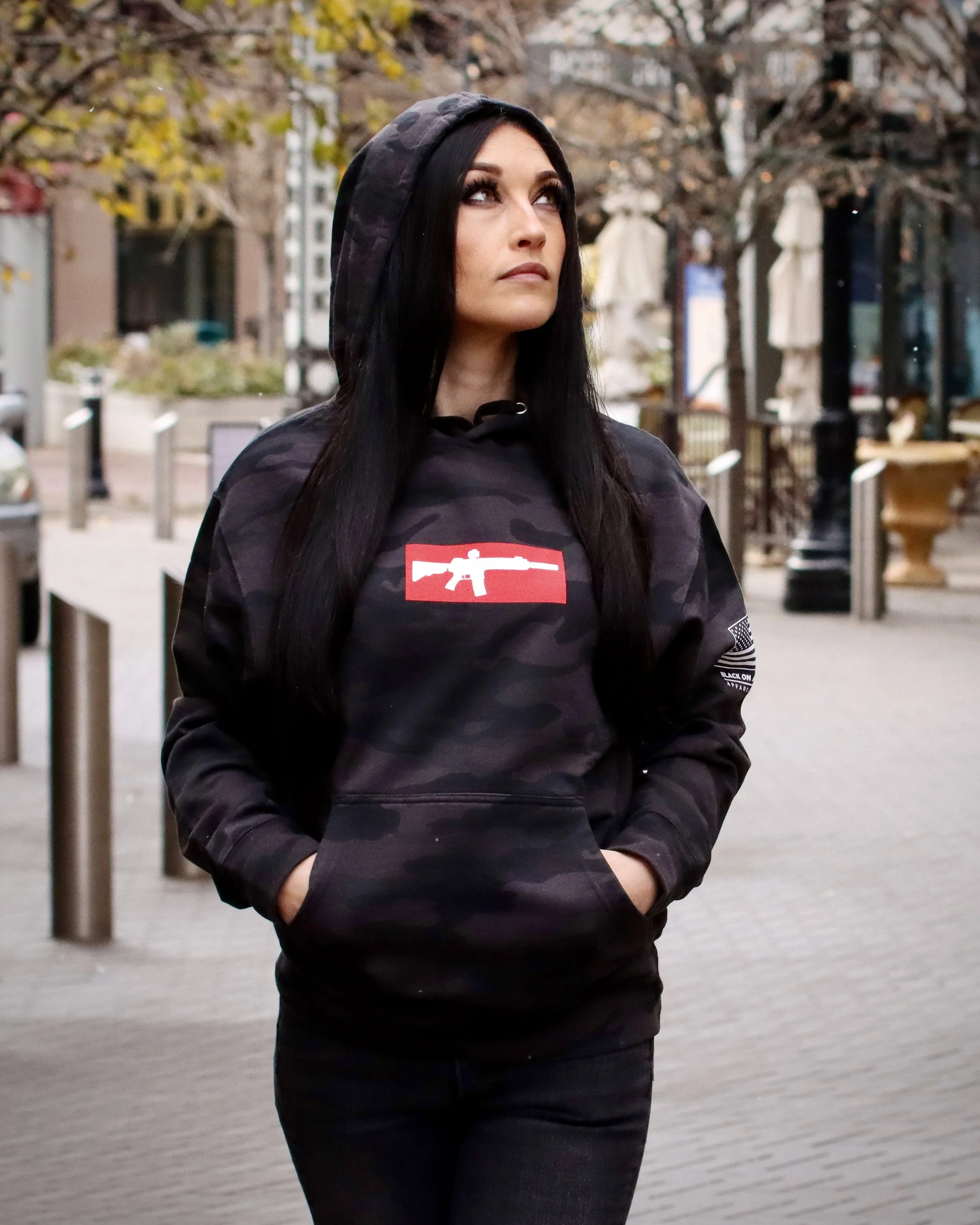 A woman with long black hair wearing a camouflaged hoodie with a red and white AR-15 graphic on the front, standing outdoors with trees and a city street in the background.