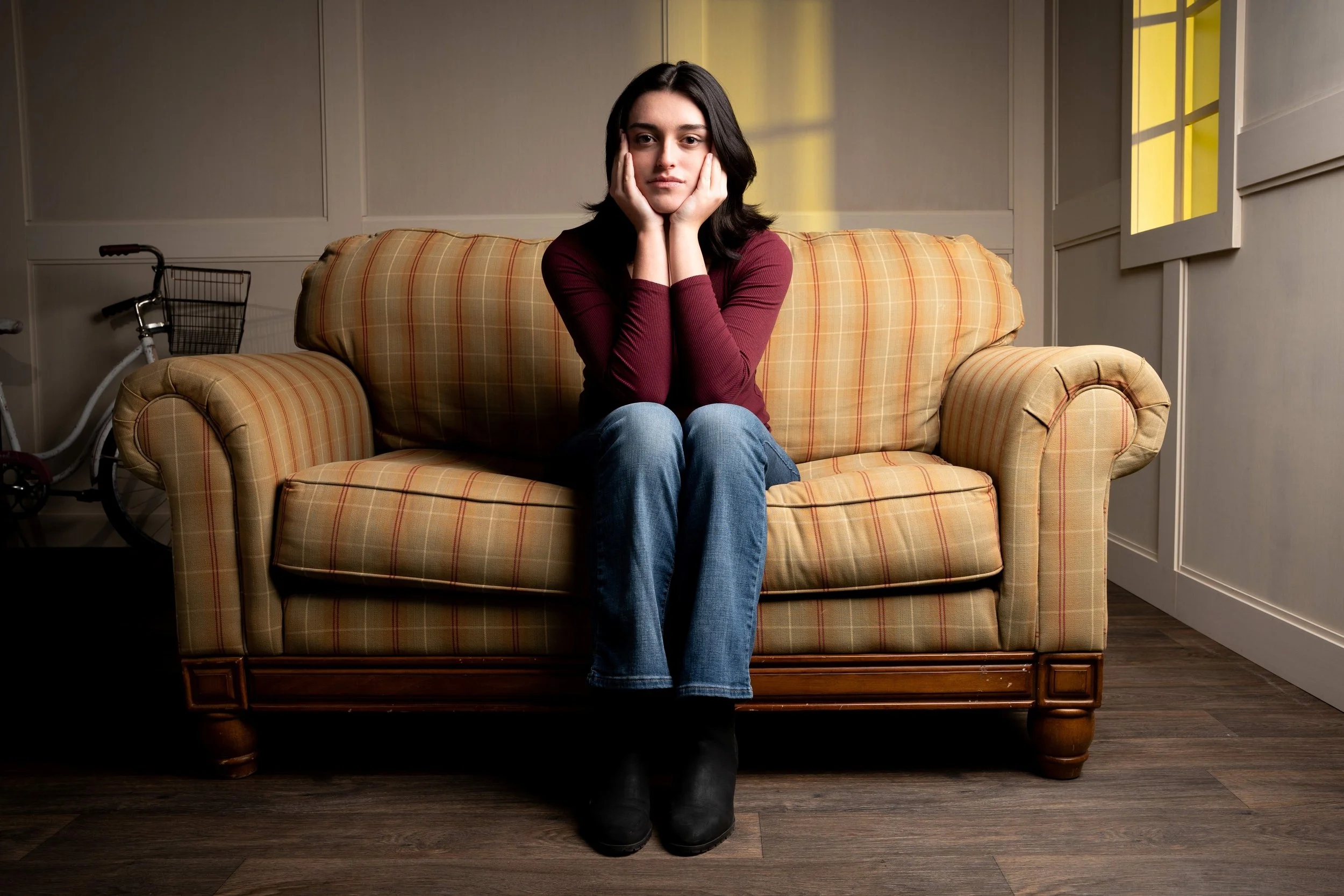 A young woman with dark hair wearing a maroon long-sleeve shirt and blue jeans sitting on a yellowish plaid sofa with her hands supporting her face, in a room with cream walls, a basket wheelchair to her left, and yellow-lit window on her right.