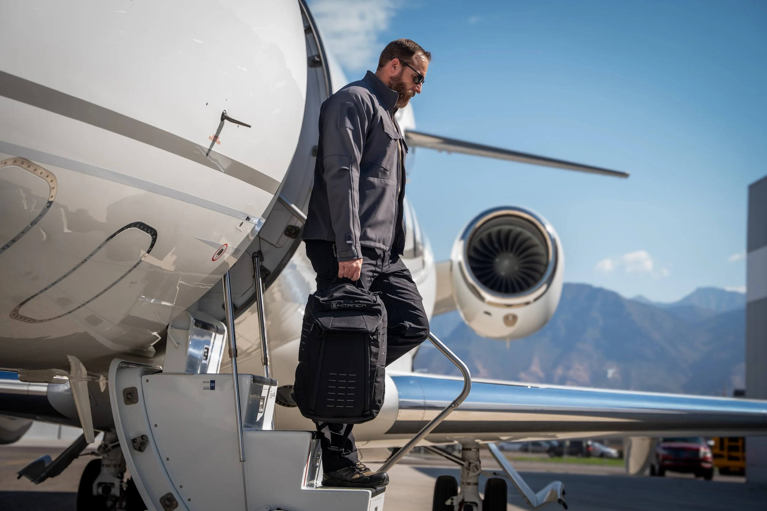 A man with a beard and sunglasses boarding a private jet with a black carry-on bag, wearing a gray jacket and black pants.