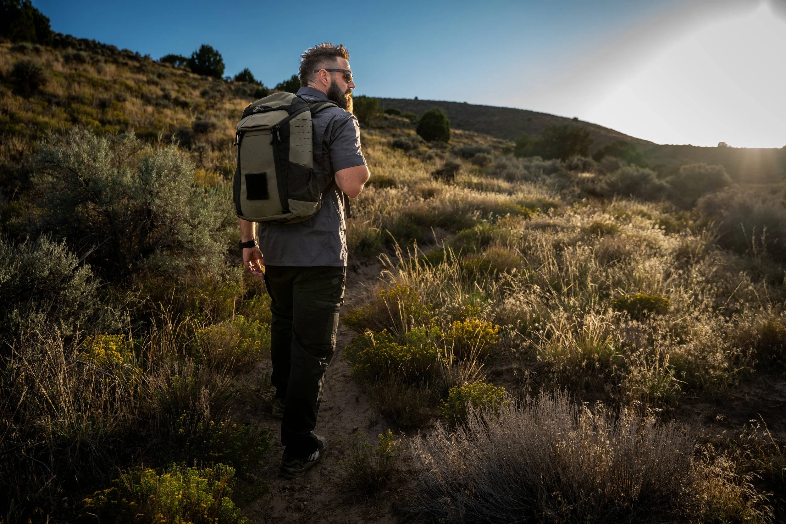 A man with a backpack and sunglasses hiking along a trail in a dry, grassy landscape with hills and sparse trees at sunset.