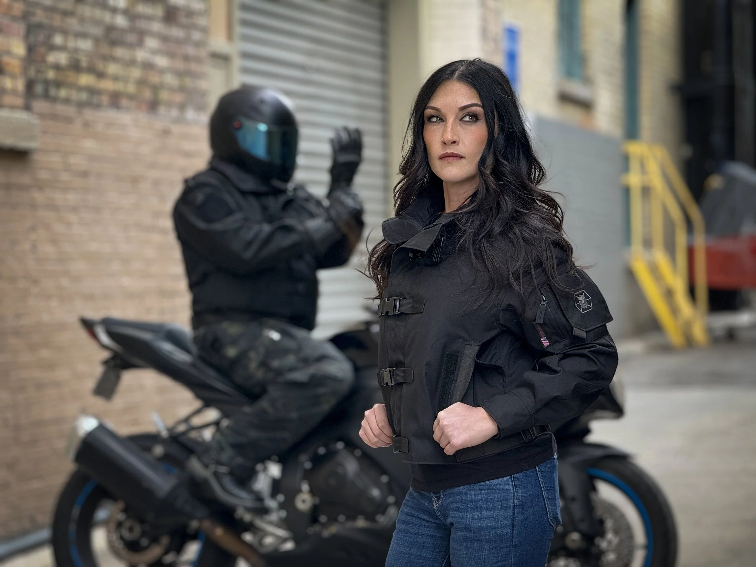 A woman with black hair and a black jacket standing outdoors near a motorbike, with an individual in motorcycle gear and helmet in the background, in an urban alleyway with brick walls.