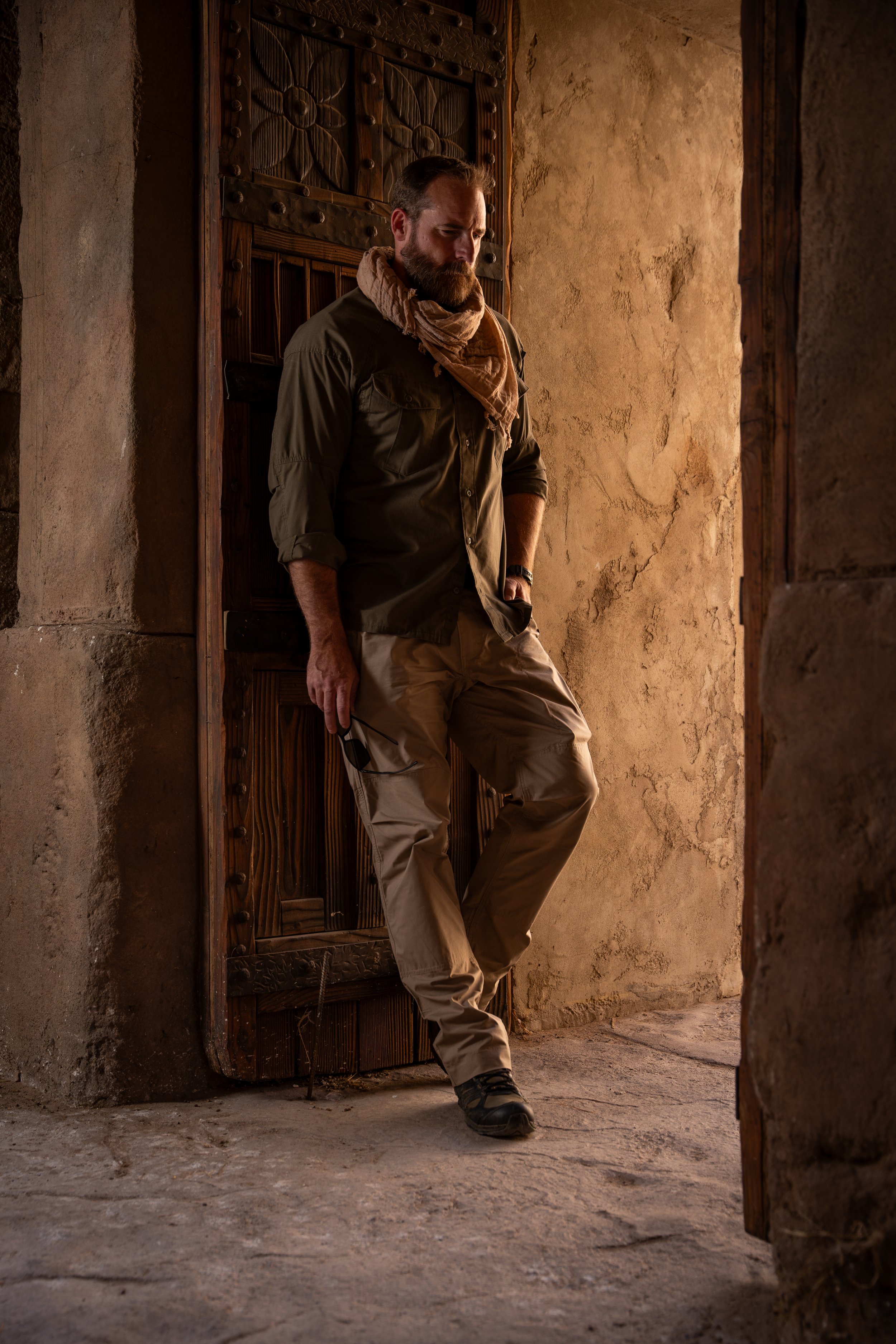A man with a beard, wearing outdoor clothing and a scarf, leaning against an ornate wooden door in a rustic, earth-toned setting.