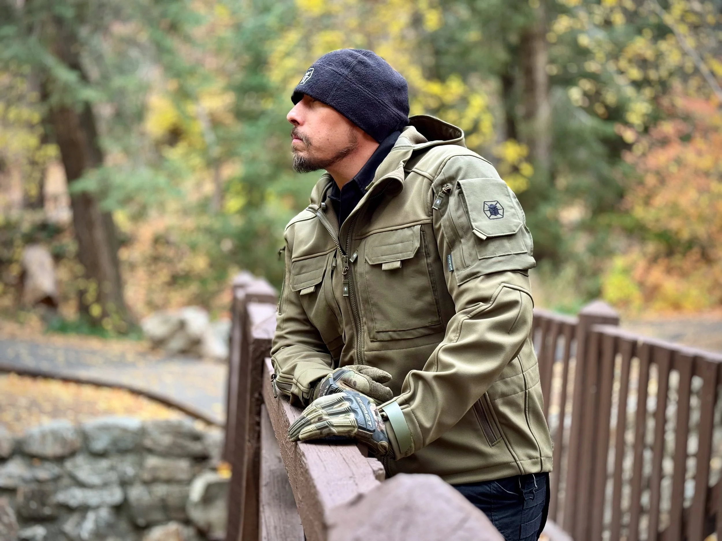 A man in a gray jacket and black beanie leaning on a park wooden railing with colorful fall foliage in the background.