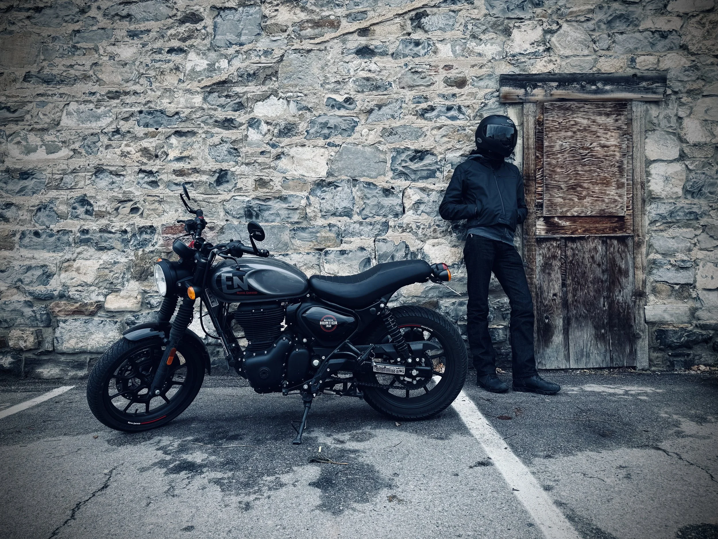 A person in motorcycle gear, wearing a black helmet and jacket, is standing against a stone wall next to a parked black motorcycle. The wall has a small boarded-up window, and the motorcycle has a label that reads 'Royal Enfield Hunter 350'.