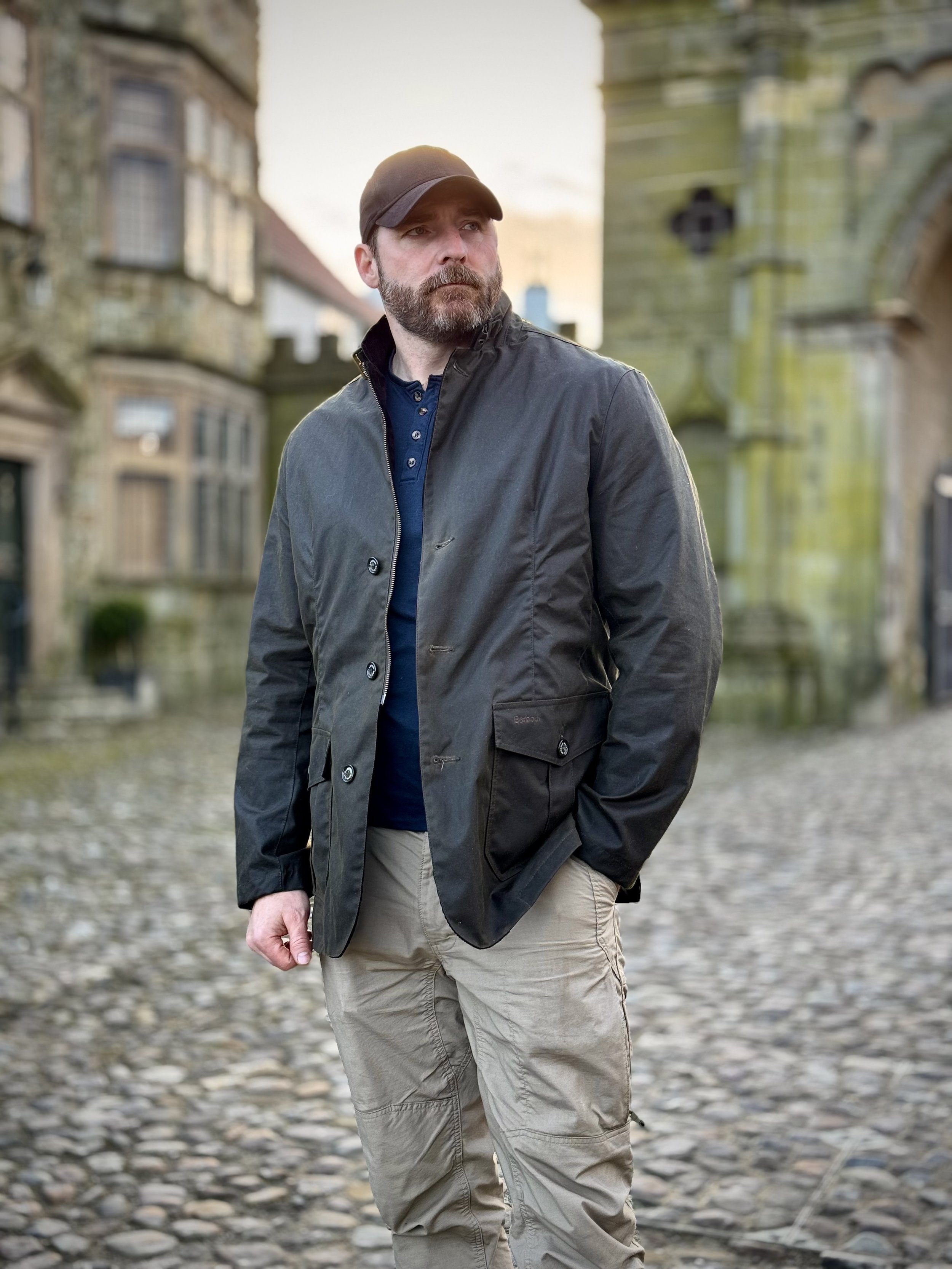 A man with a beard and mustache wearing a black baseball cap, black jacket, and beige cargo pants standing outdoors on a cobblestone street with old buildings and lampposts in the background during daylight.