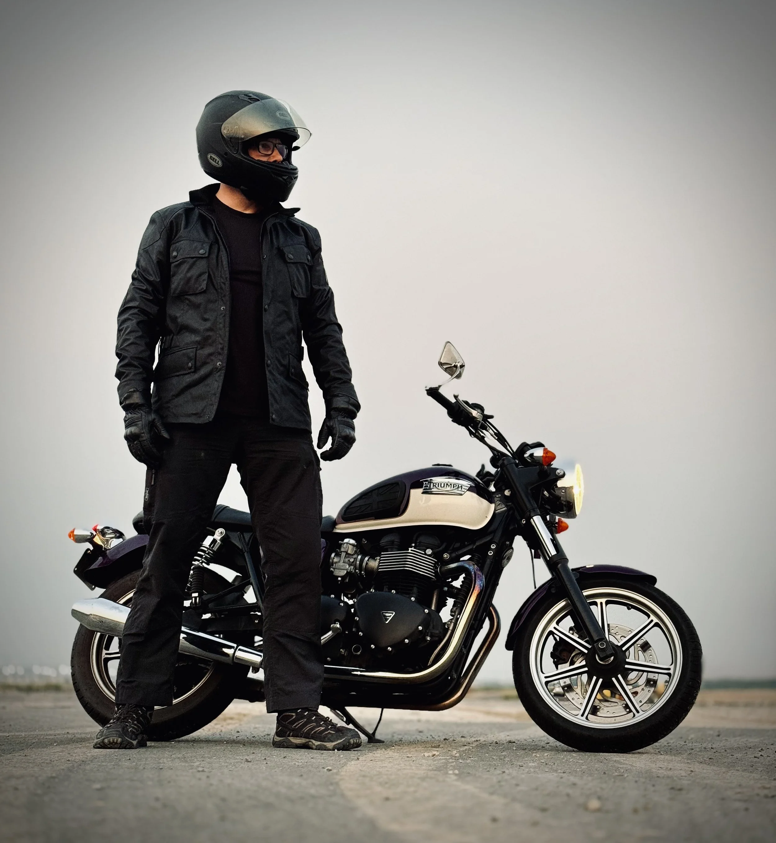Person wearing a full-face motorcycle helmet, black jacket, black gloves, and black pants, standing beside a black and white Triumph motorcycle on an open ground with a cloudy sky in the background.