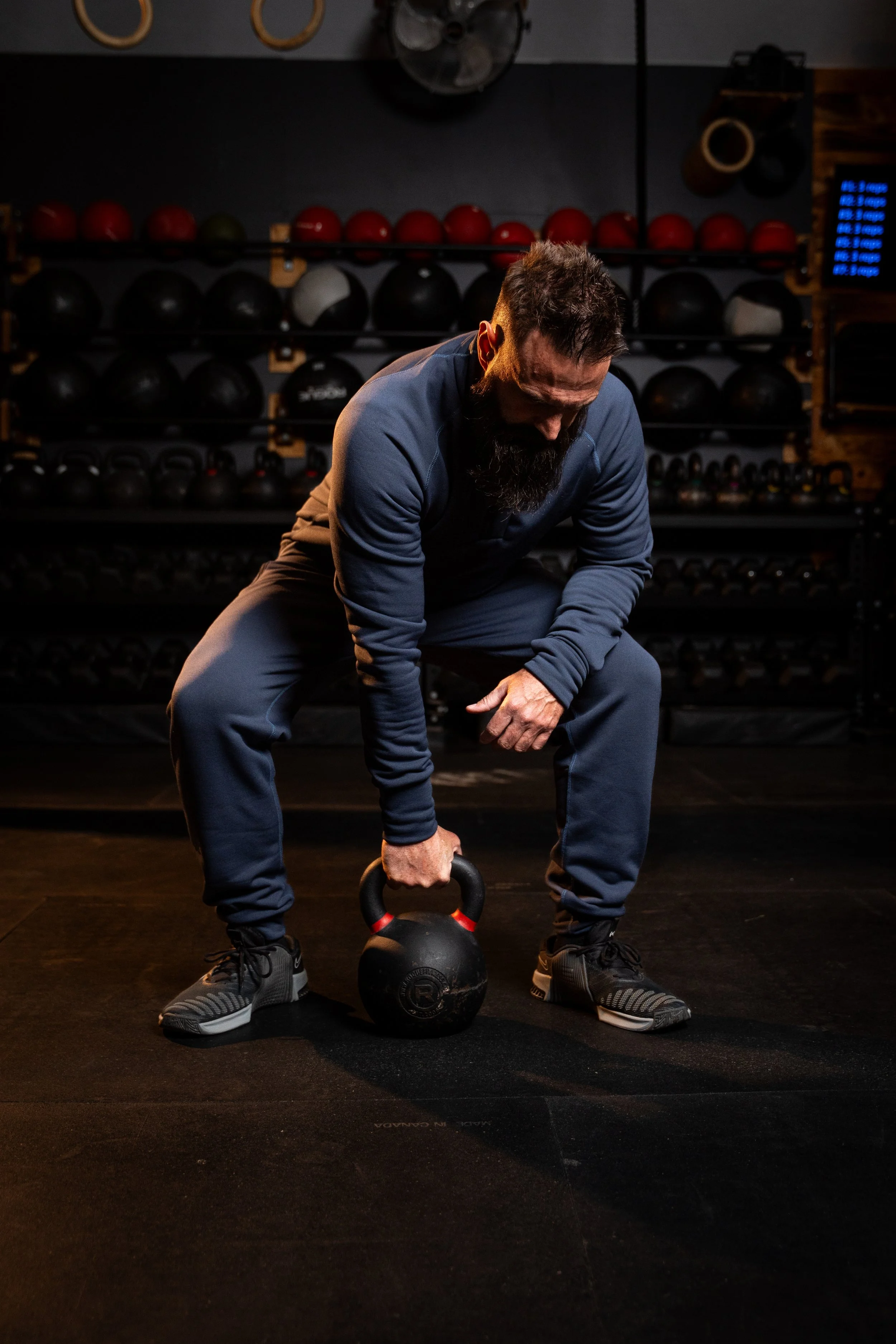 A man with a beard and workout clothes is working out in a gym. He is bent over picking up a kettlebell, with a rack of medicine balls and kettlebells in the background.